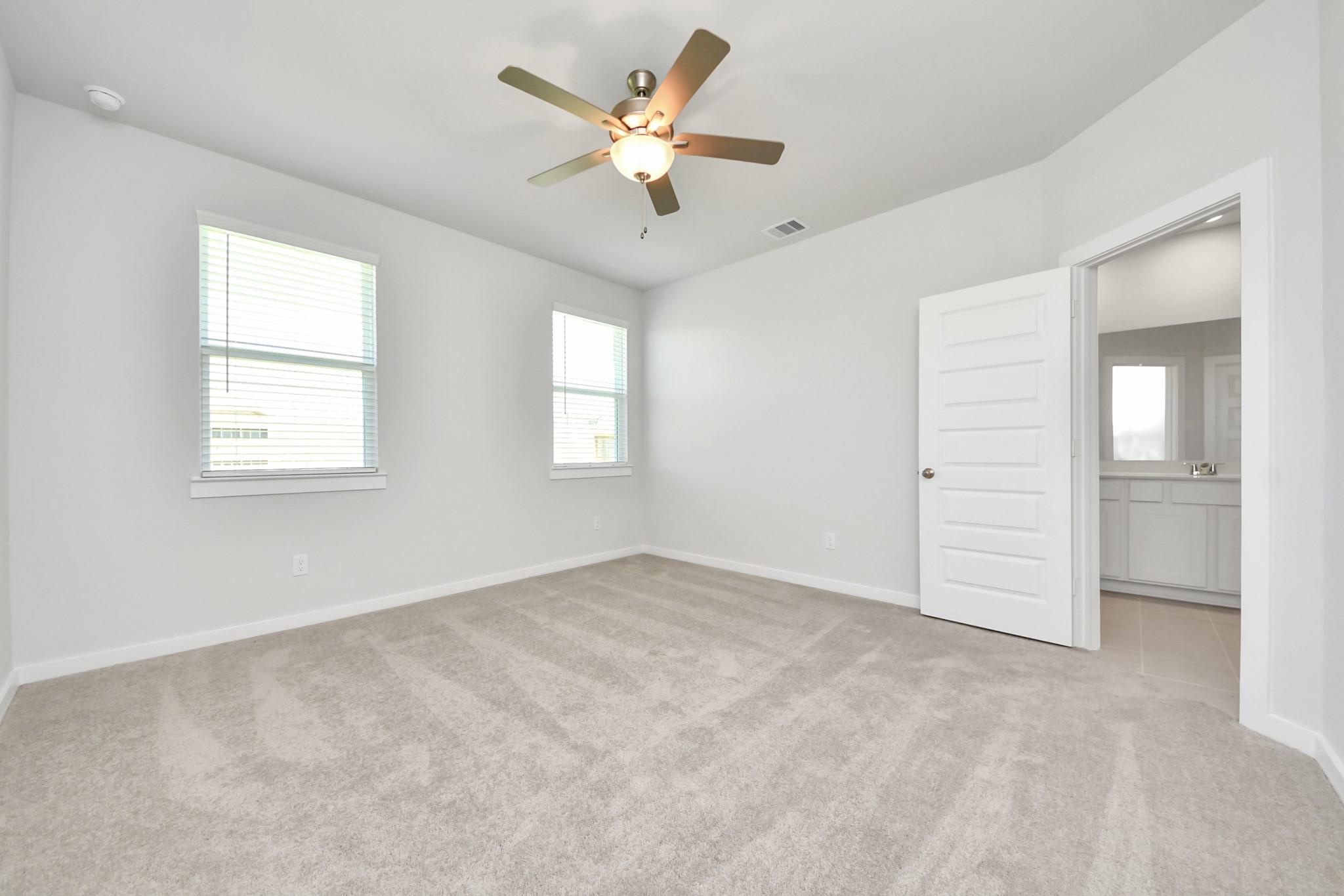 Bright carpeted bedroom with ceiling fan, two windows, and en-suite bath access in Davidson Homes The Costa A, Emberly, Beasley TX