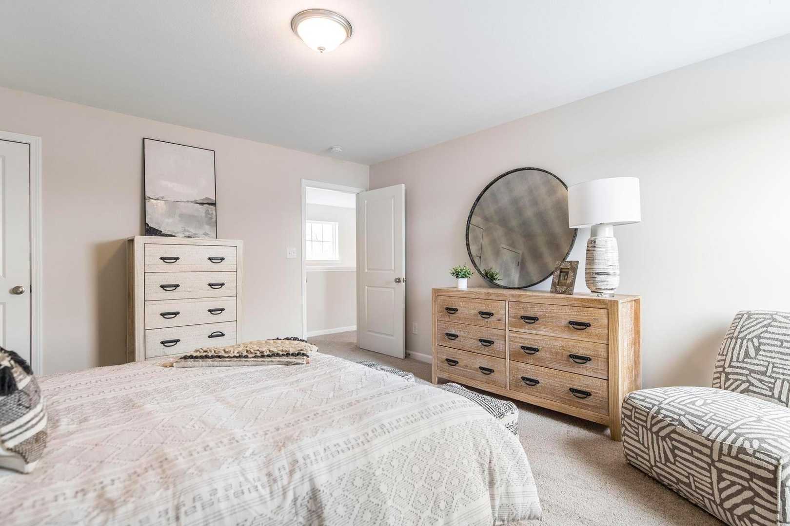 Spacious primary bedroom at Cape Reserve Donahue Ridge in Auburn AL with neutral bedding, wooden dressers, round mirror and armchair