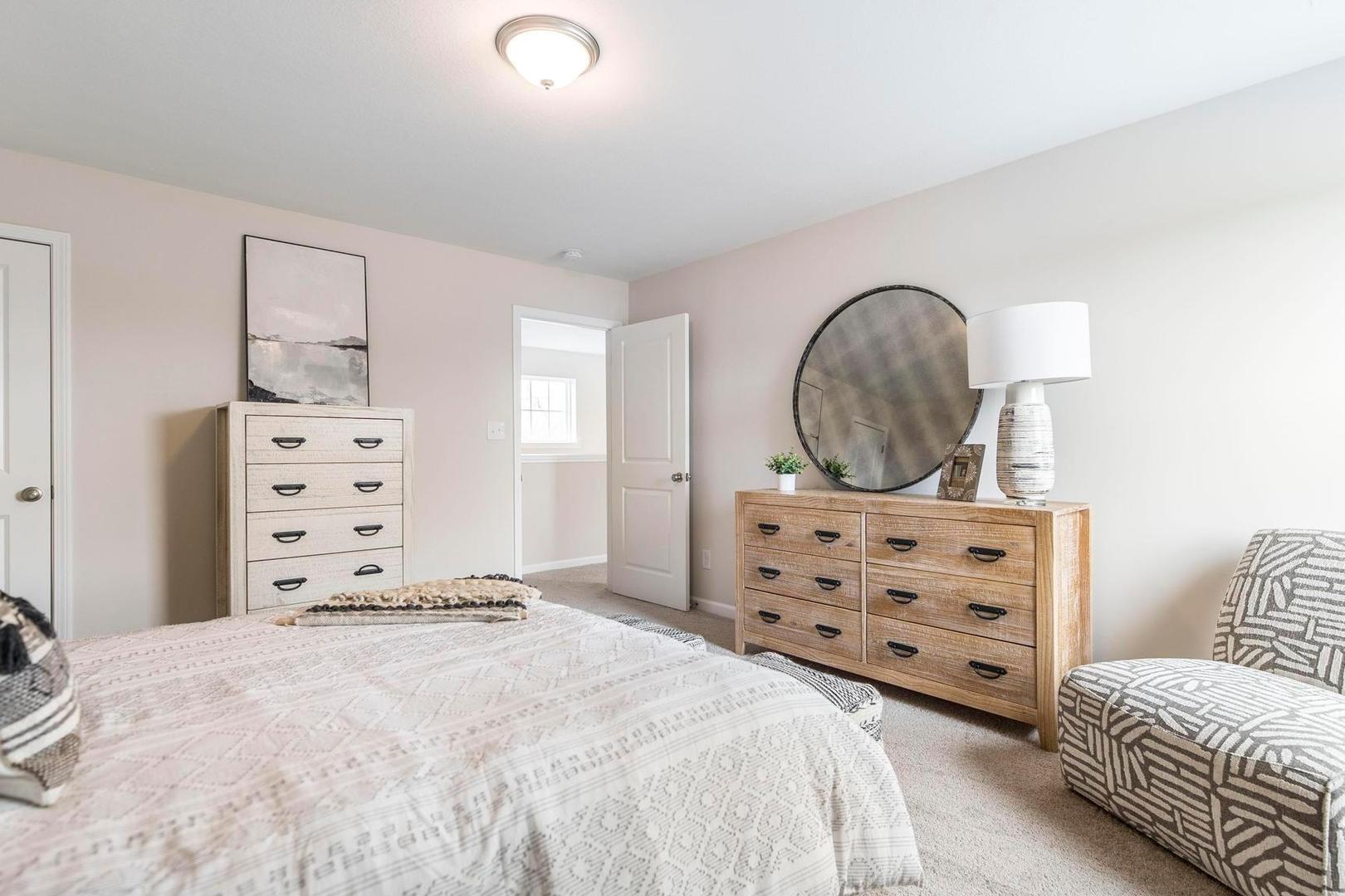 Spacious primary bedroom at Cape Reserve Donahue Ridge in Auburn AL with neutral bedding, wooden dressers, round mirror and armchair