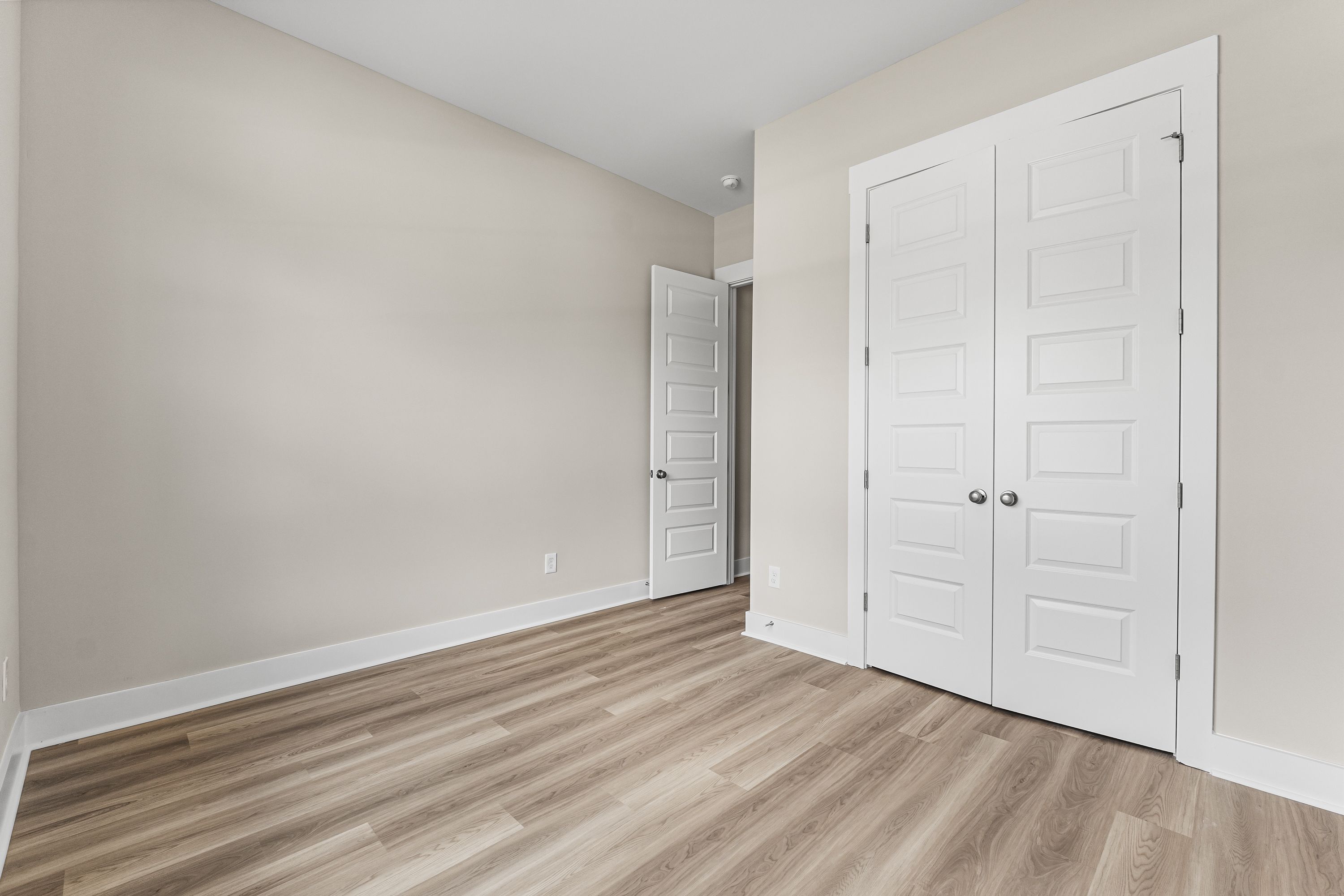 Spacious empty bedroom in The Oxford A home with beige walls, white bi-fold closet doors, open entry door, and light hardwood floors