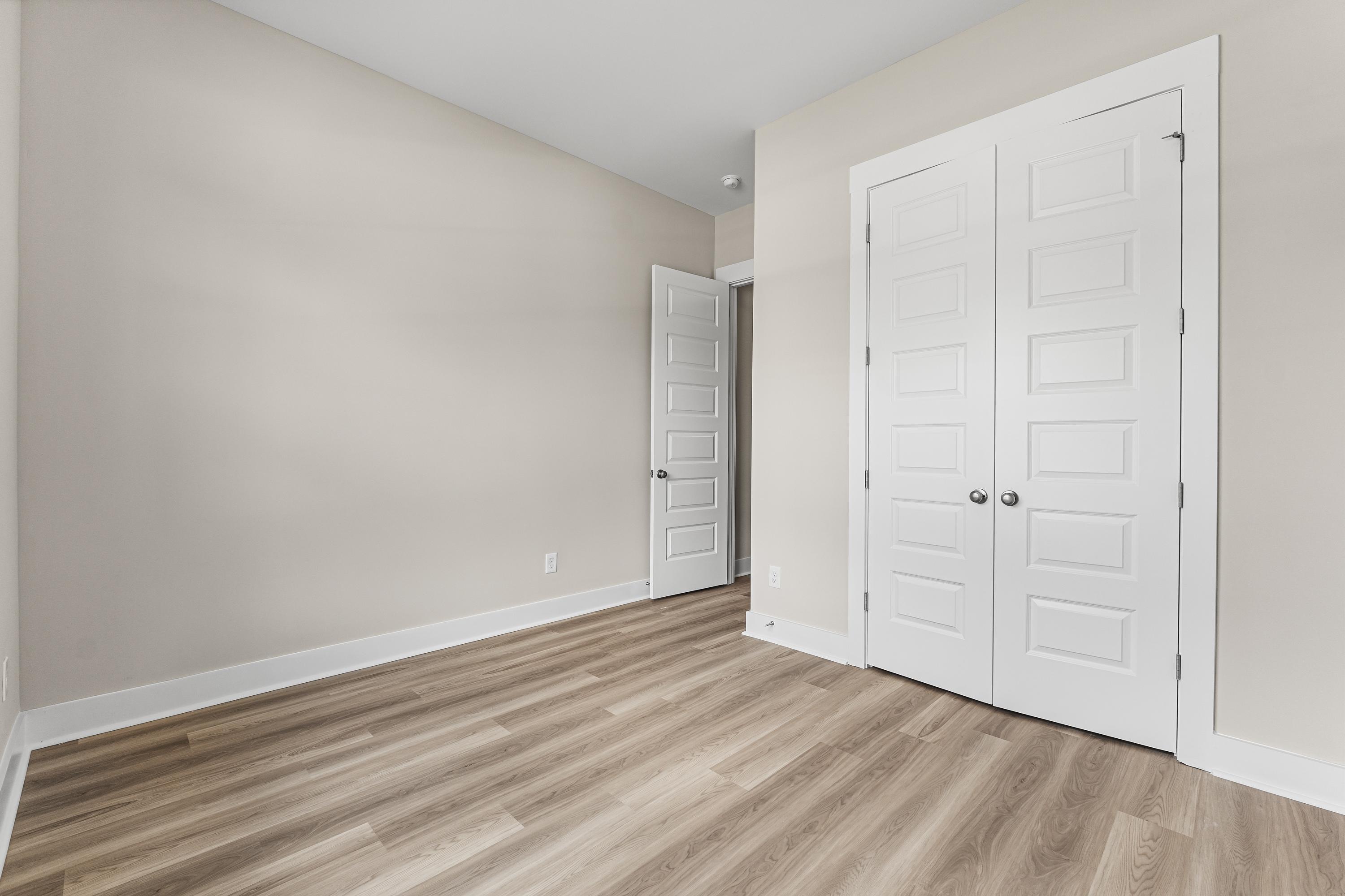 Spacious empty bedroom in The Oxford A home with beige walls, white bi-fold closet doors, open entry door, and light hardwood floors