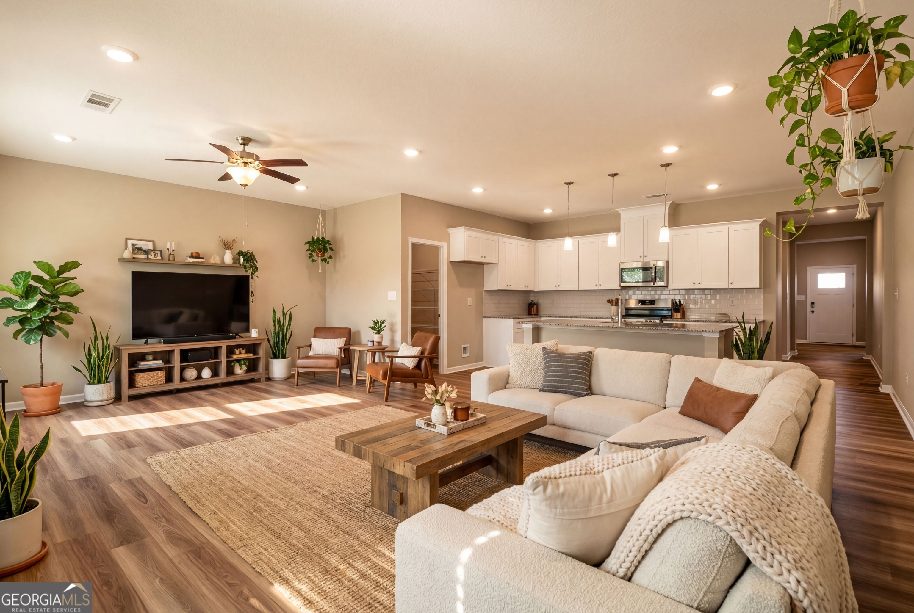 Open-concept living room with beige sofa, wooden coffee table, TV, and potted plants next to white kitchen in Evermore Homes The Luna, Perry, Georgia