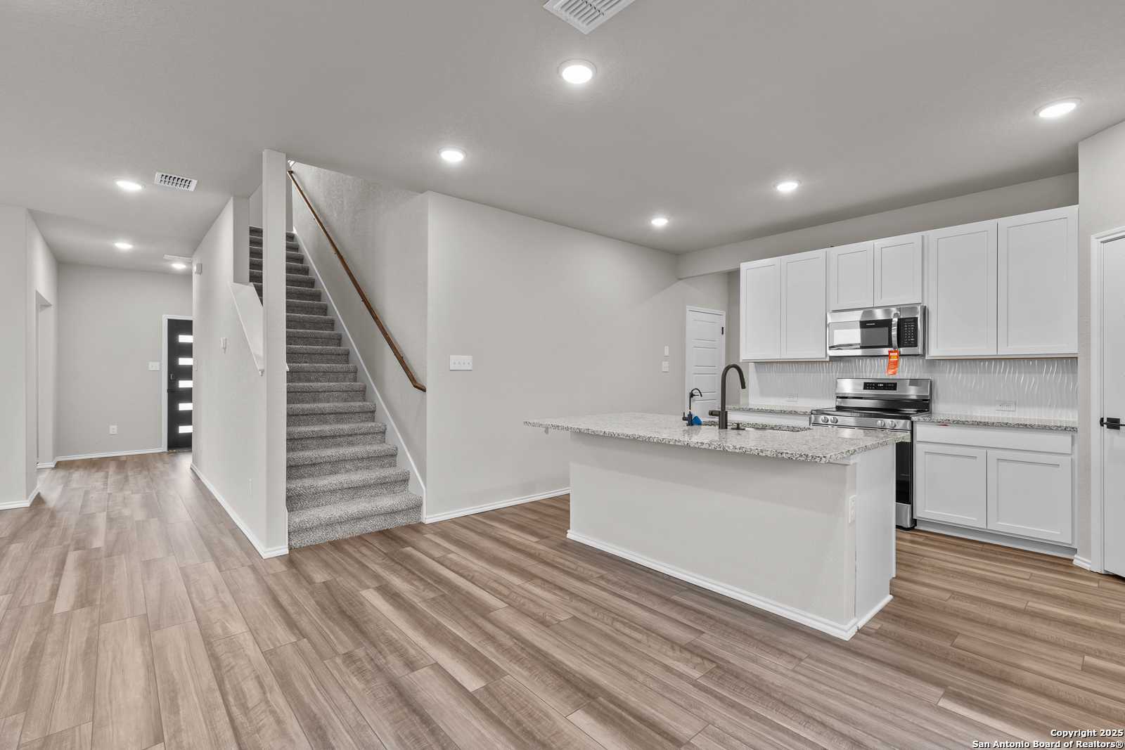 Open kitchen with white cabinets, granite island, stainless appliances, and carpeted staircase in The Murray I 4-bedroom home, San Antonio