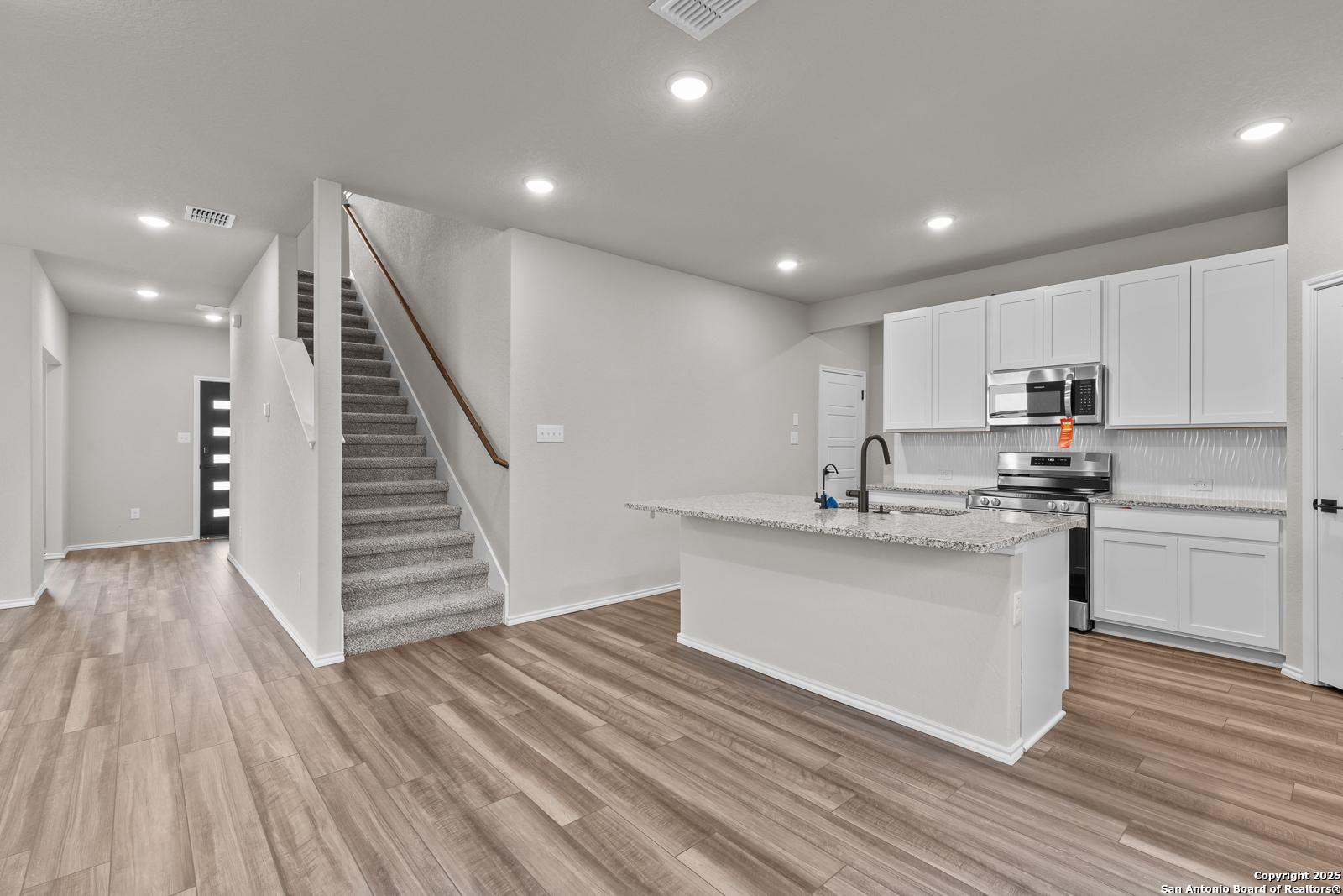 Open kitchen with white cabinets, granite island, stainless appliances, and carpeted staircase in The Murray I 4-bedroom home, San Antonio