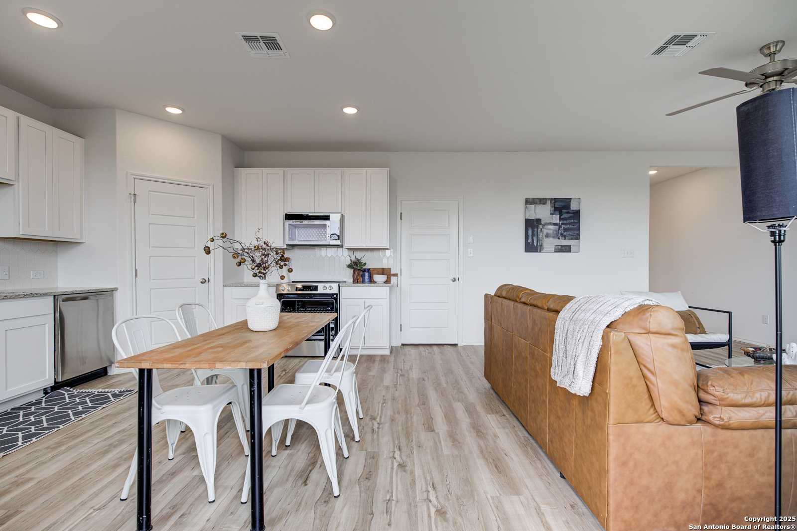 Open-concept living and dining area with white kitchen cabinets, wooden table, tan leather sofa in Davidson Homes The Trinity A, San Antonio