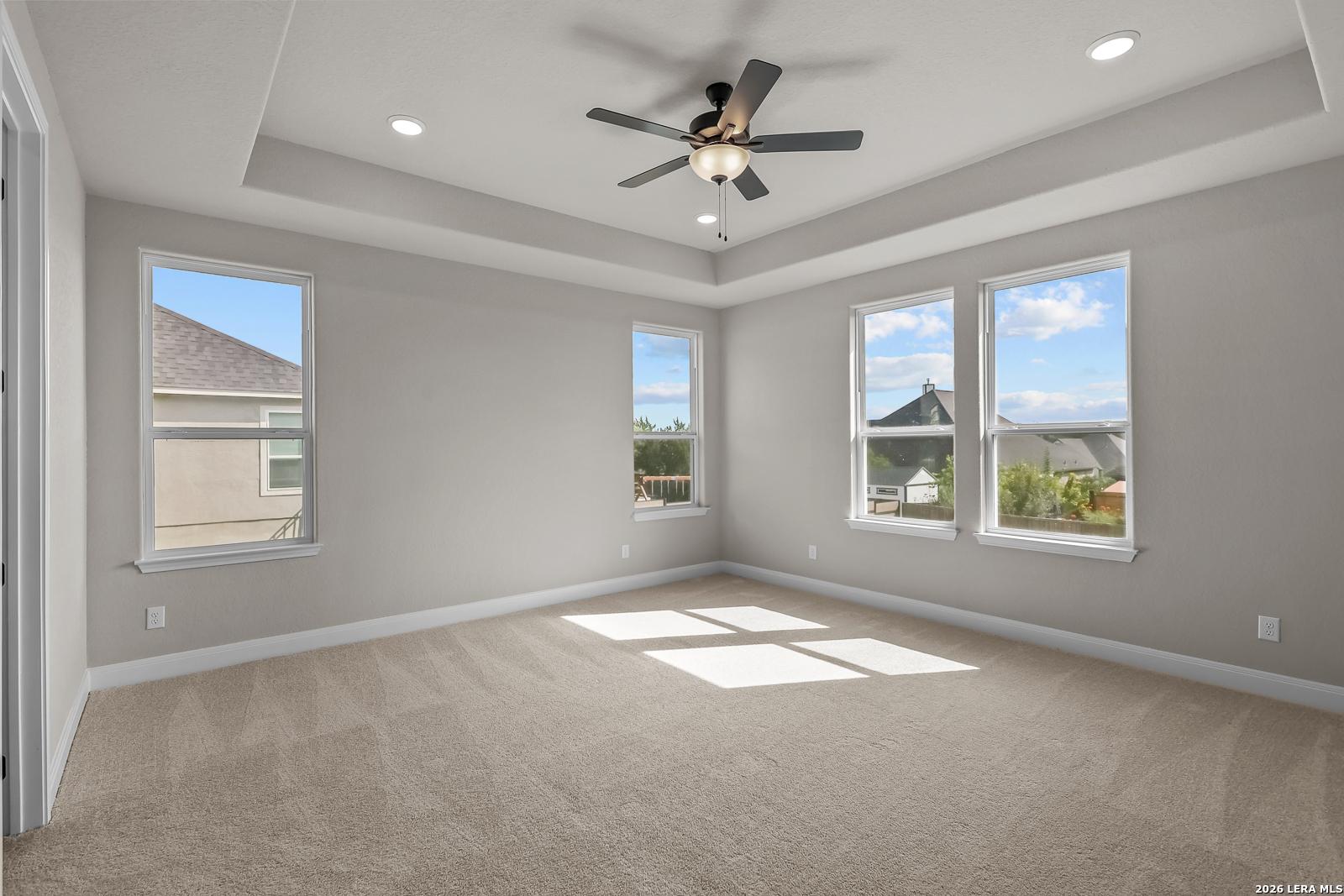 Bright bedroom with tray ceiling, ceiling fan, and multiple windows in Davidson Homes The Foster E, Castroville, Texas