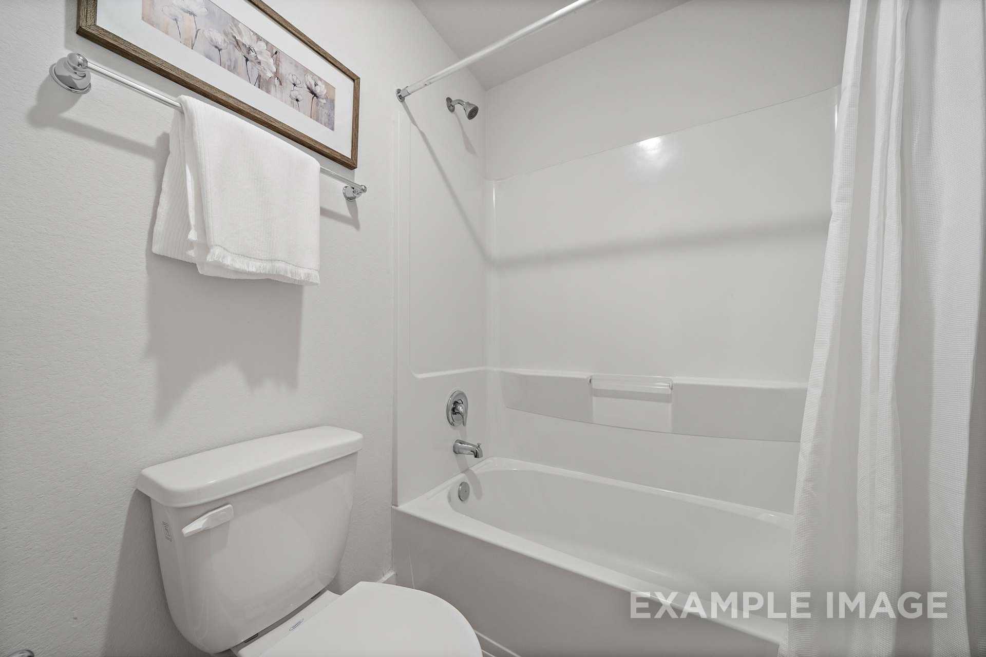 Bright white bathroom in The Rose B home design featuring tub-shower combo, toilet, towels, and wall art