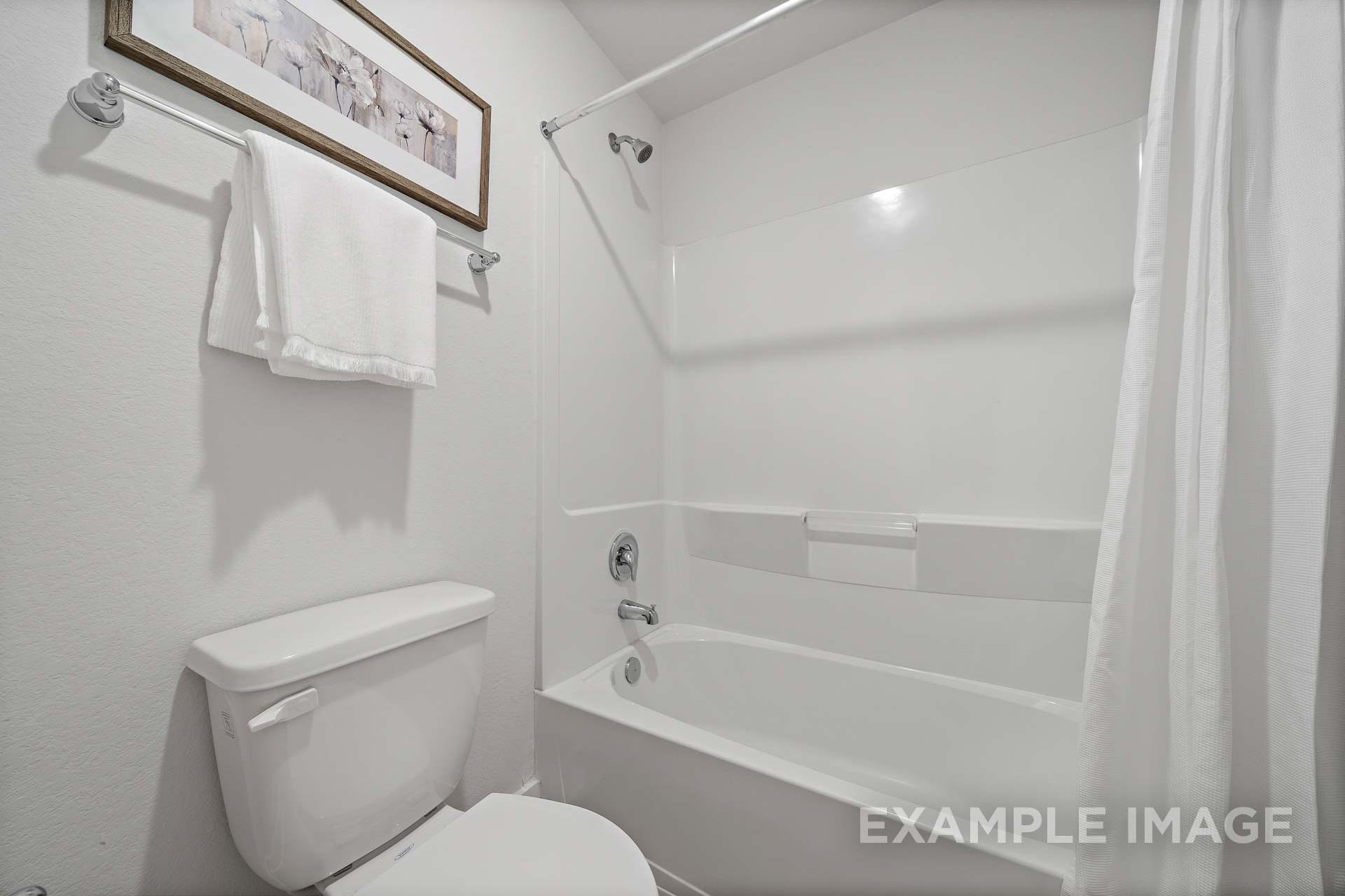 Bright white bathroom in The Rose B home design featuring tub-shower combo, toilet, towels, and wall art