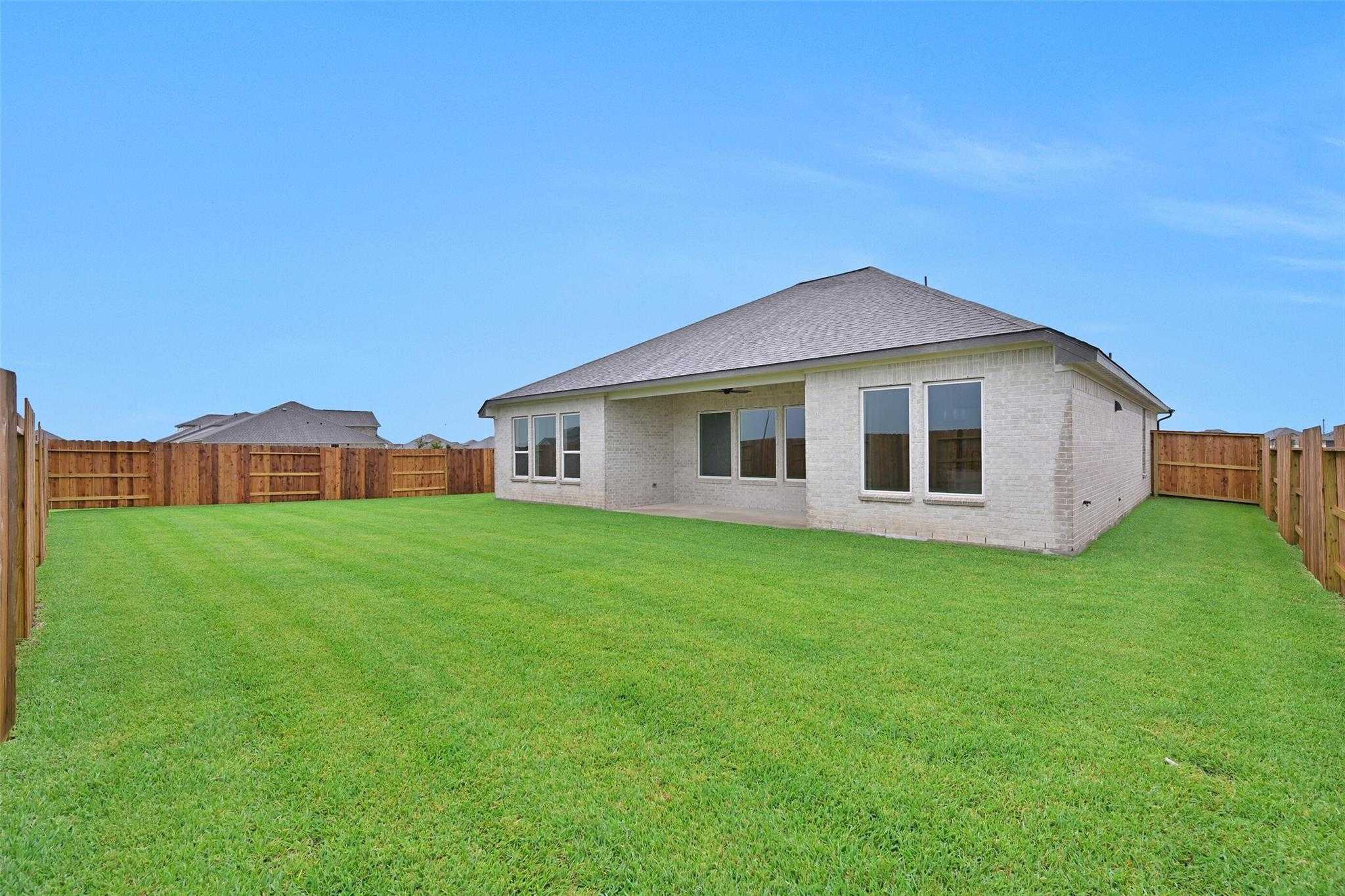 Rear view of brick single-story home with covered patio, large green yard, and wooden fences in Lago Mar, Texas City
