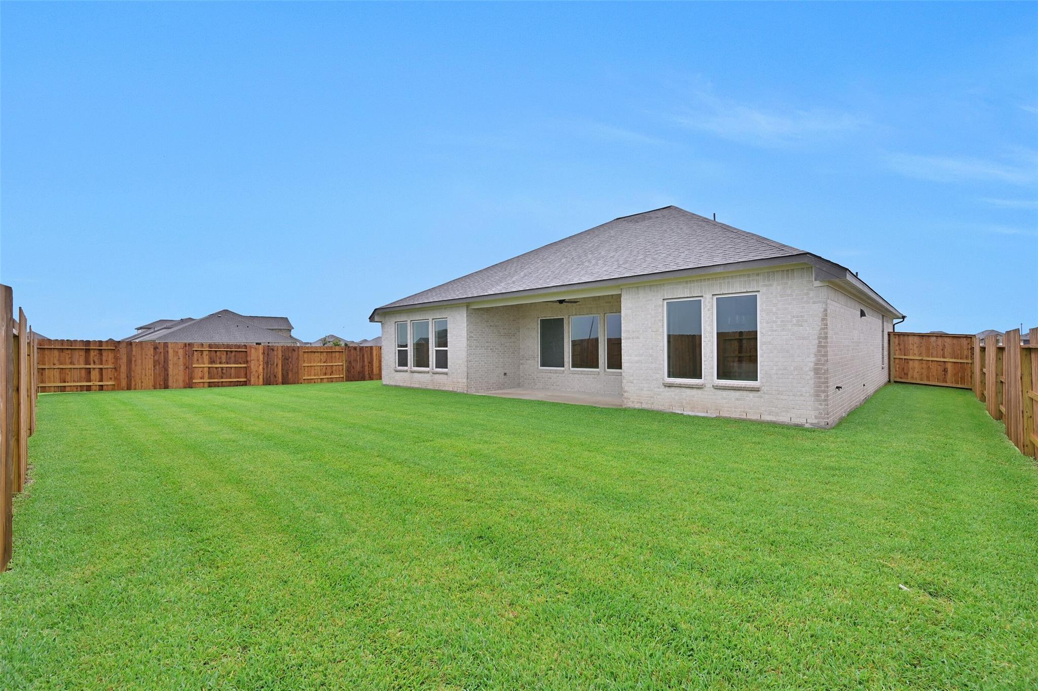 Rear view of brick single-story home with covered patio, large green yard, and wooden fences in Lago Mar, Texas City