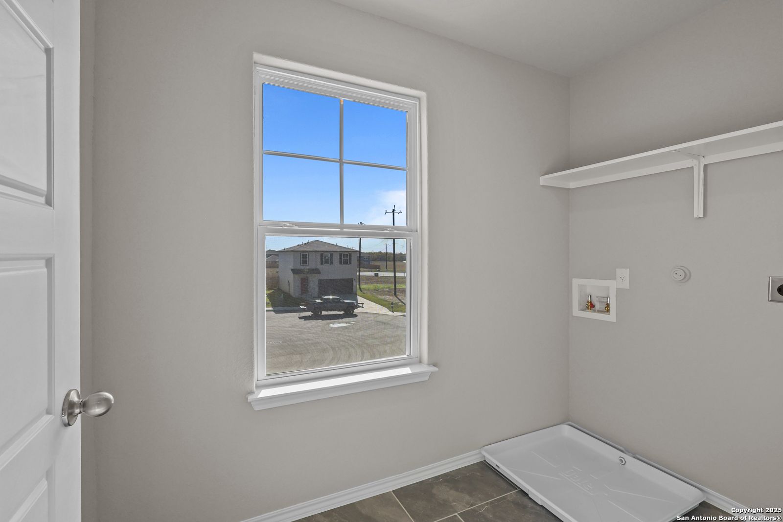 Bright laundry room with large window overlooking driveway, built-in shelves, and washer-dryer hookups in 4-bedroom Trinity D home, San Antonio
