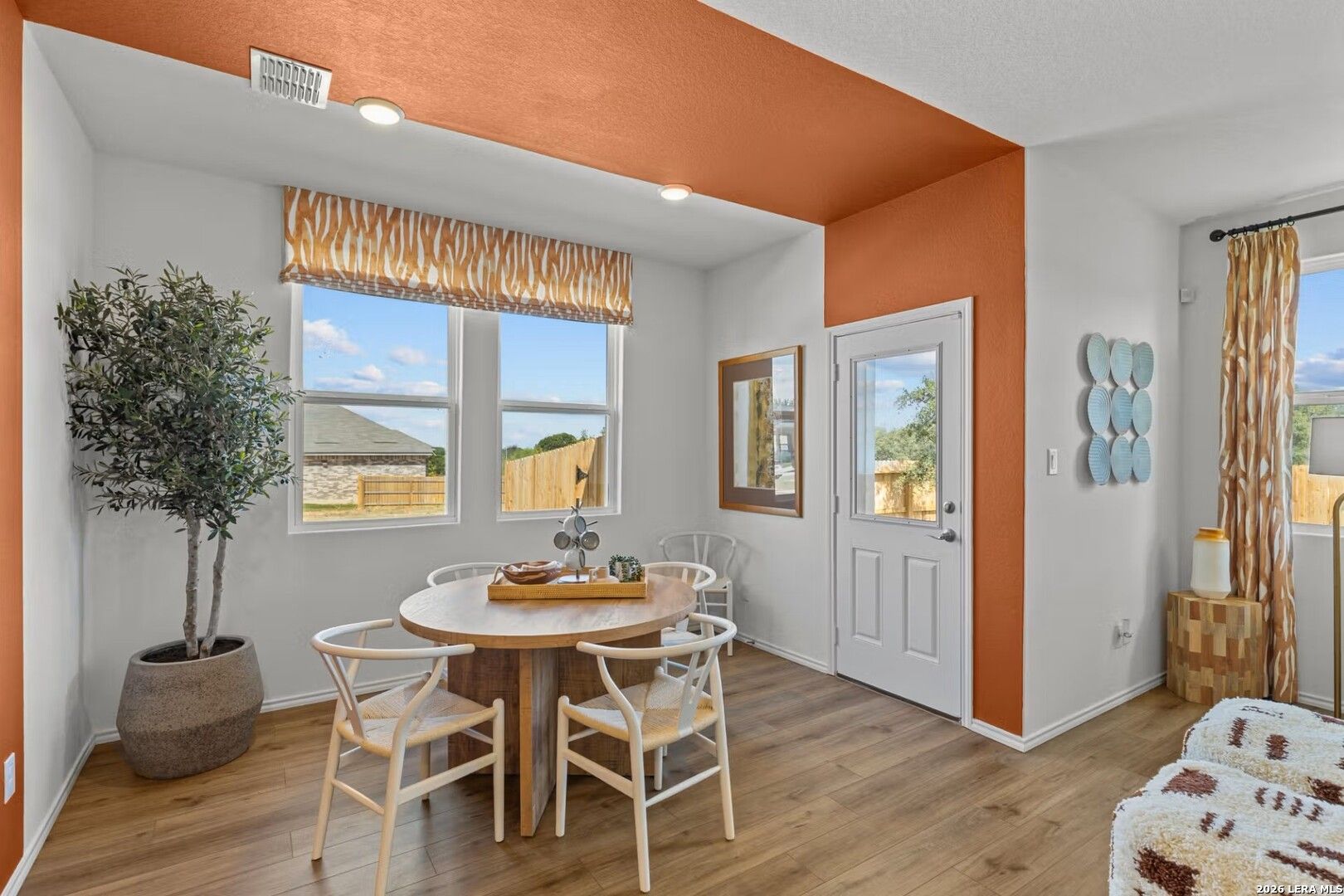 Cozy breakfast nook with round wood table, white wishbone chairs, potted olive tree, orange accent wall in Davidson Homes The Daphne K, Bricewood San Antonio