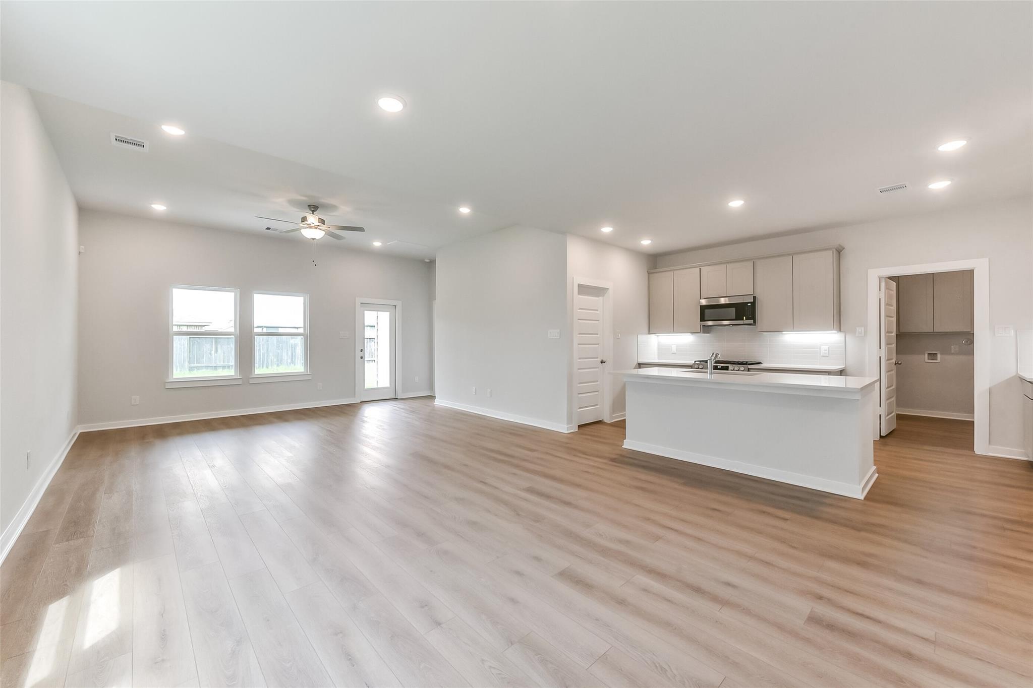 Spacious open living room with light hardwood floors, ceiling fan, large windows, and modern white kitchen island in The Tierra C home, Dayton Texas