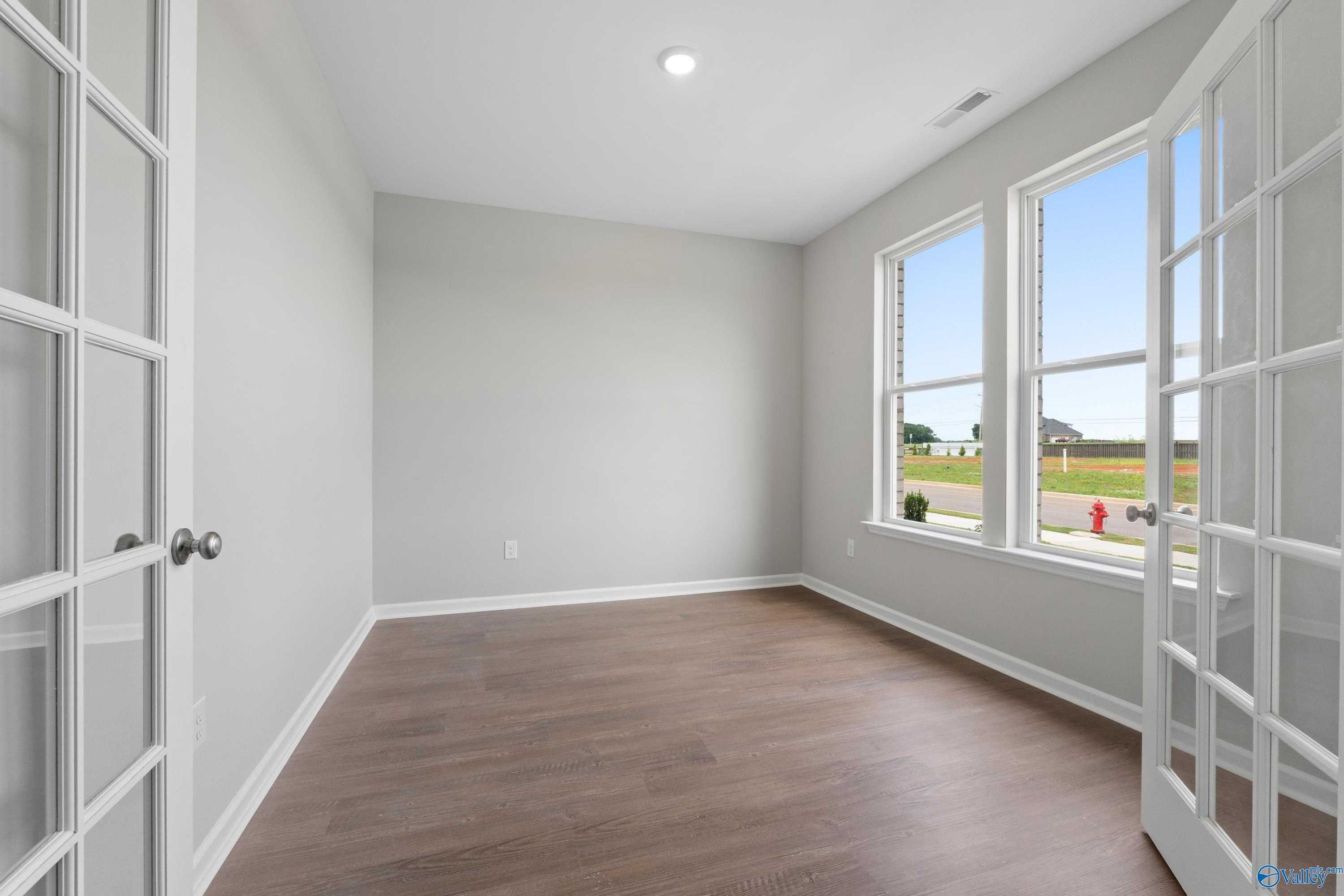 Spacious empty room with light gray walls, hardwood floors, French doors, and large neighborhood-view windows in Davidson Homes The Dorado, Athens, Alabama