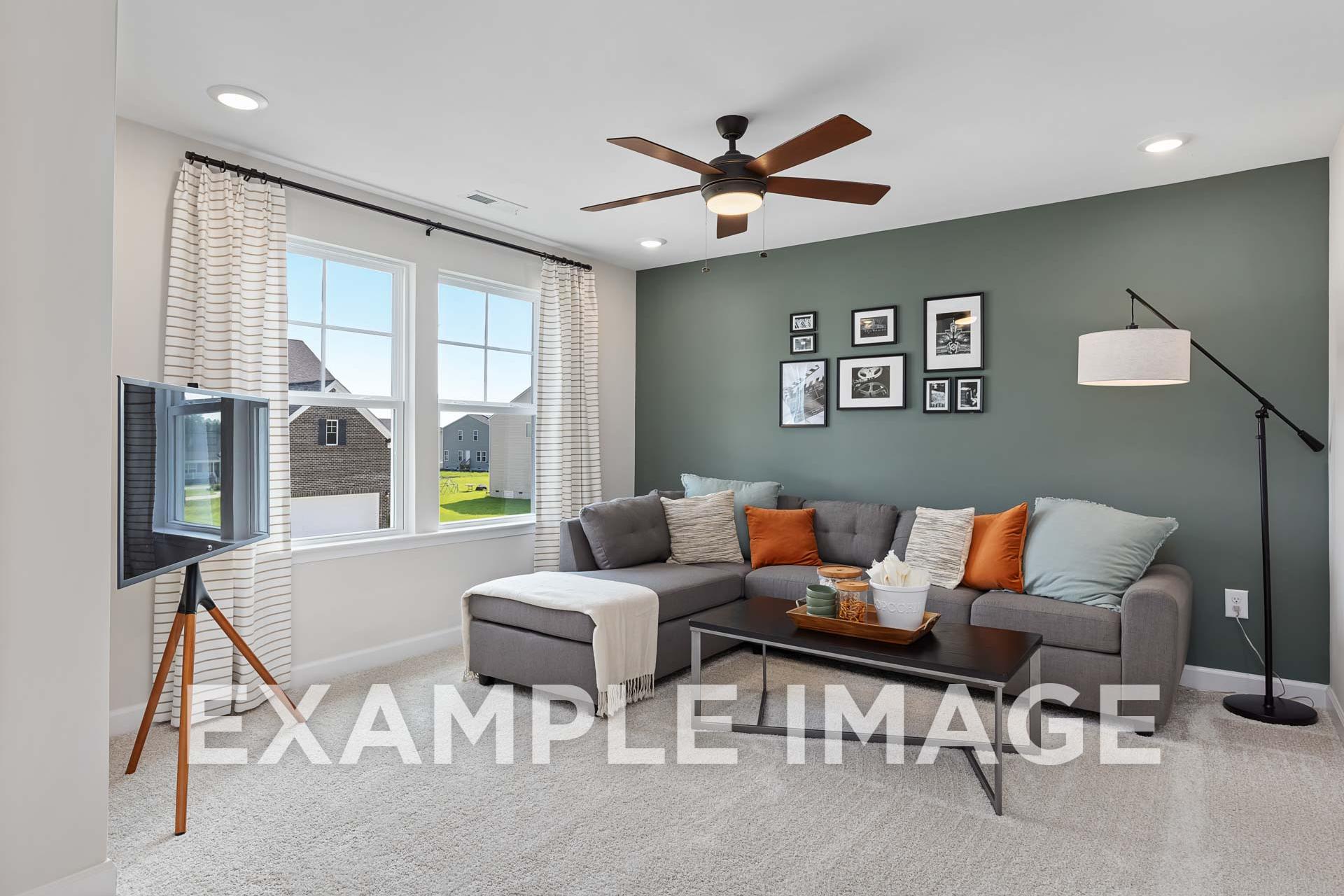 Cozy upper-floor bonus room in The Willow E with L-shaped gray sofa, orange pillows, green accent wall, TV, and large windows overlooking yard