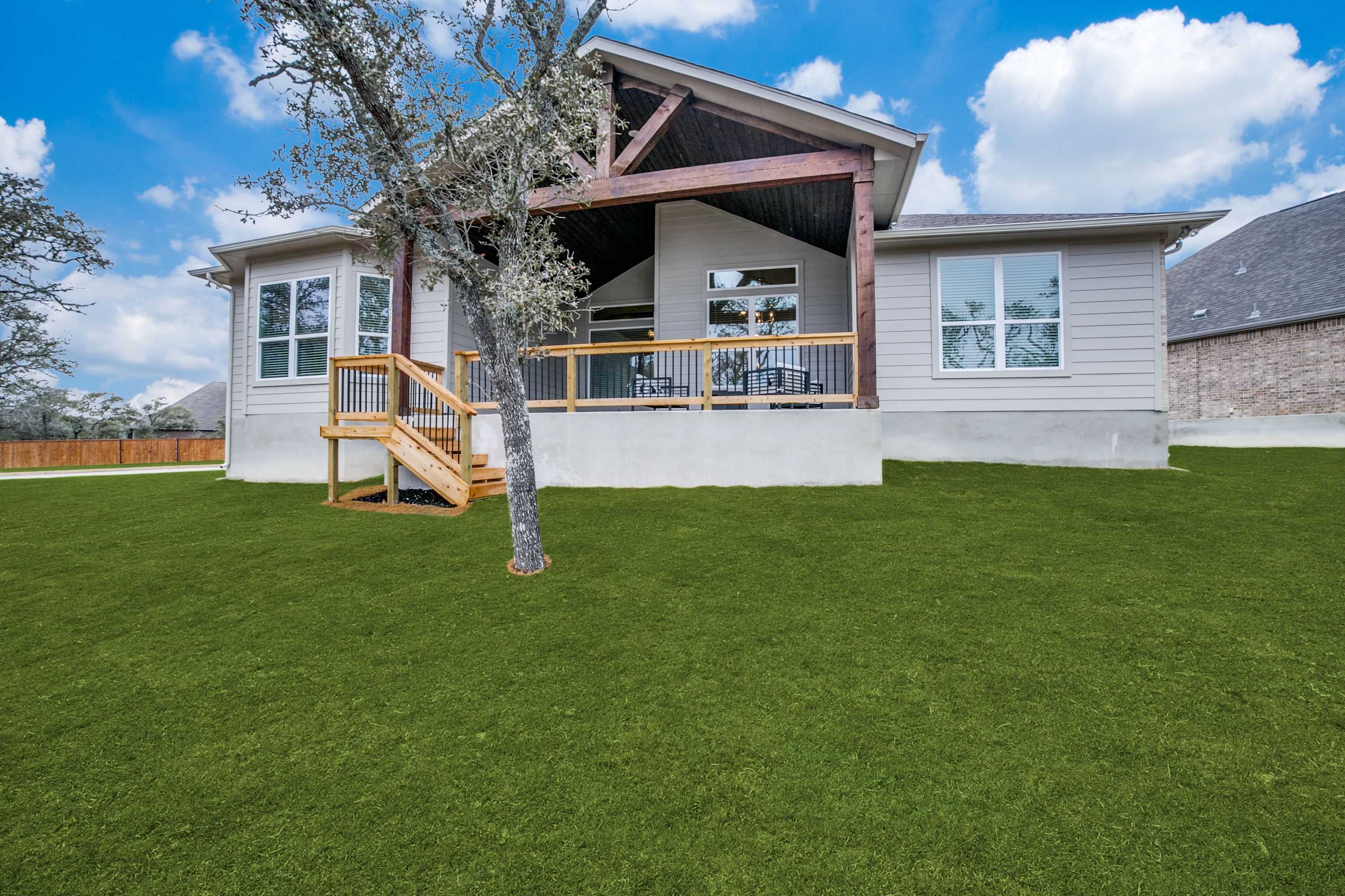 Rear elevation of The Garner A single-story home with covered patio, wooden beams, stairs, and lush green lawn in San Antonio
