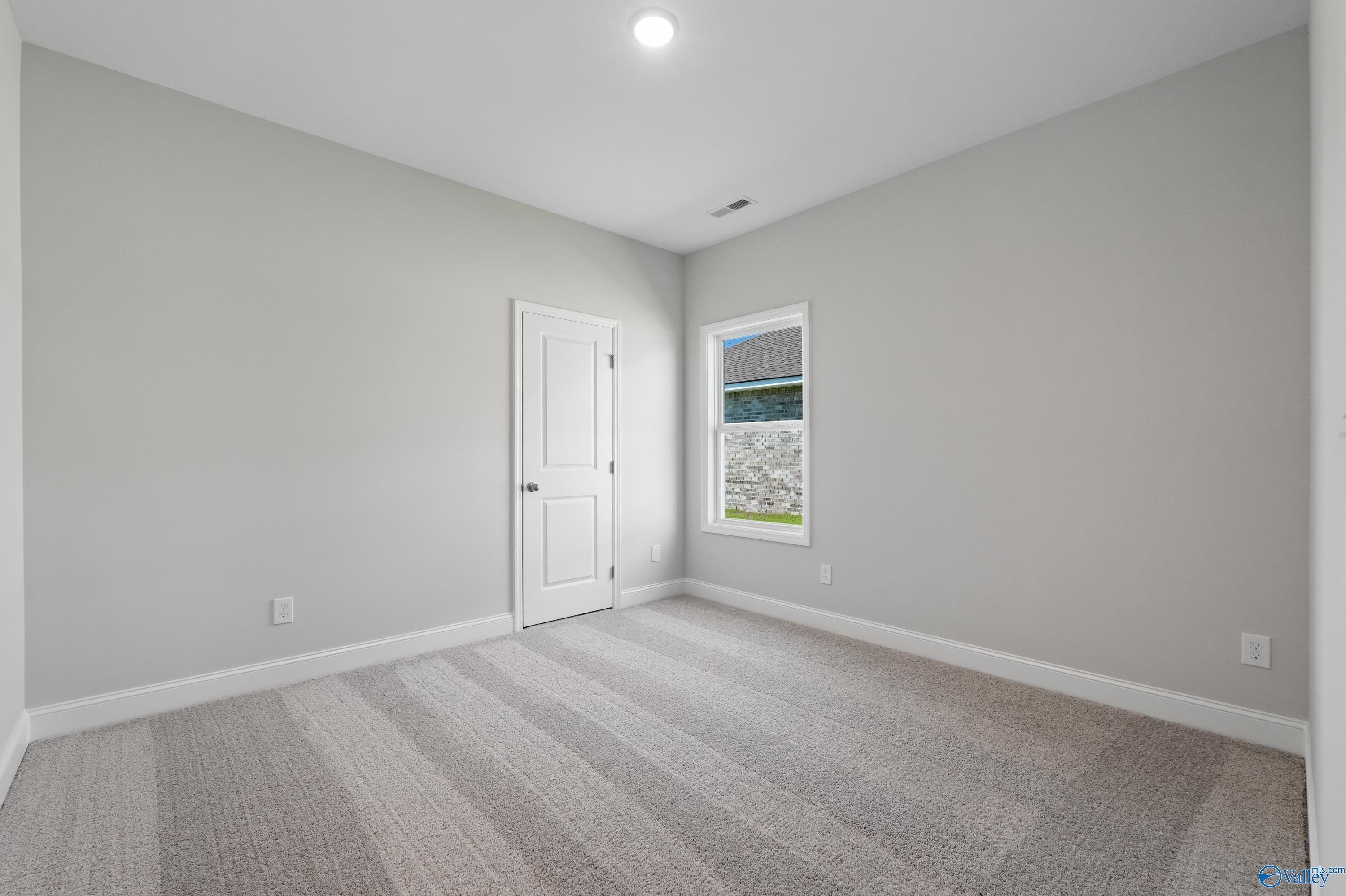 Spacious empty bedroom with neutral gray walls, striped carpet, white door, and window in Davidson Homes The Everett, Hazel Green, AL