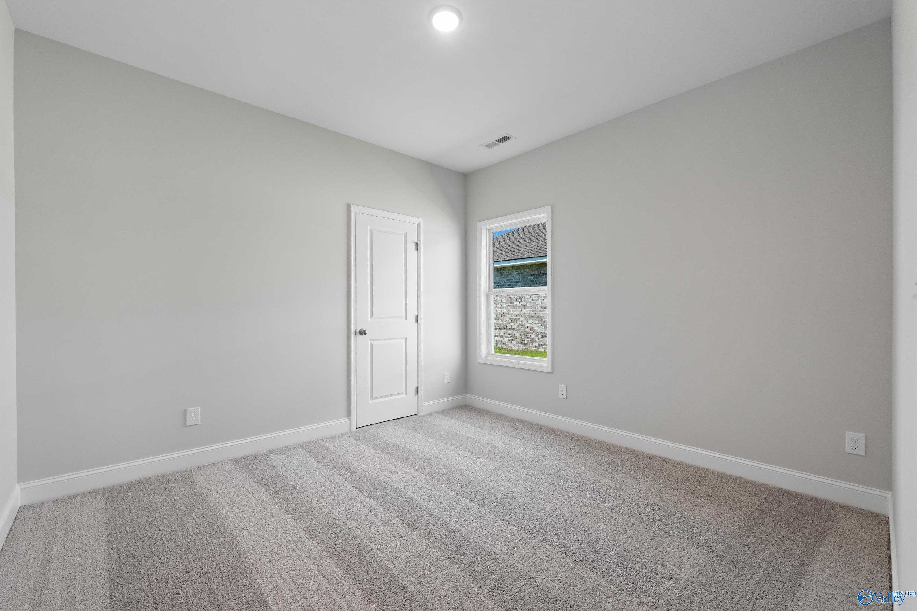 Spacious empty bedroom with neutral gray walls, striped carpet, white door, and window in Davidson Homes The Everett, Hazel Green, AL