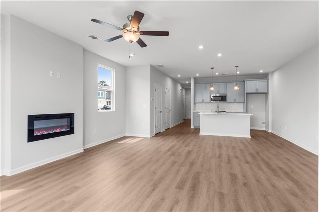 Open living room with electric fireplace, ceiling fan, hardwood floors, and adjacent white kitchen island in The Monroe B home, Emerson, GA