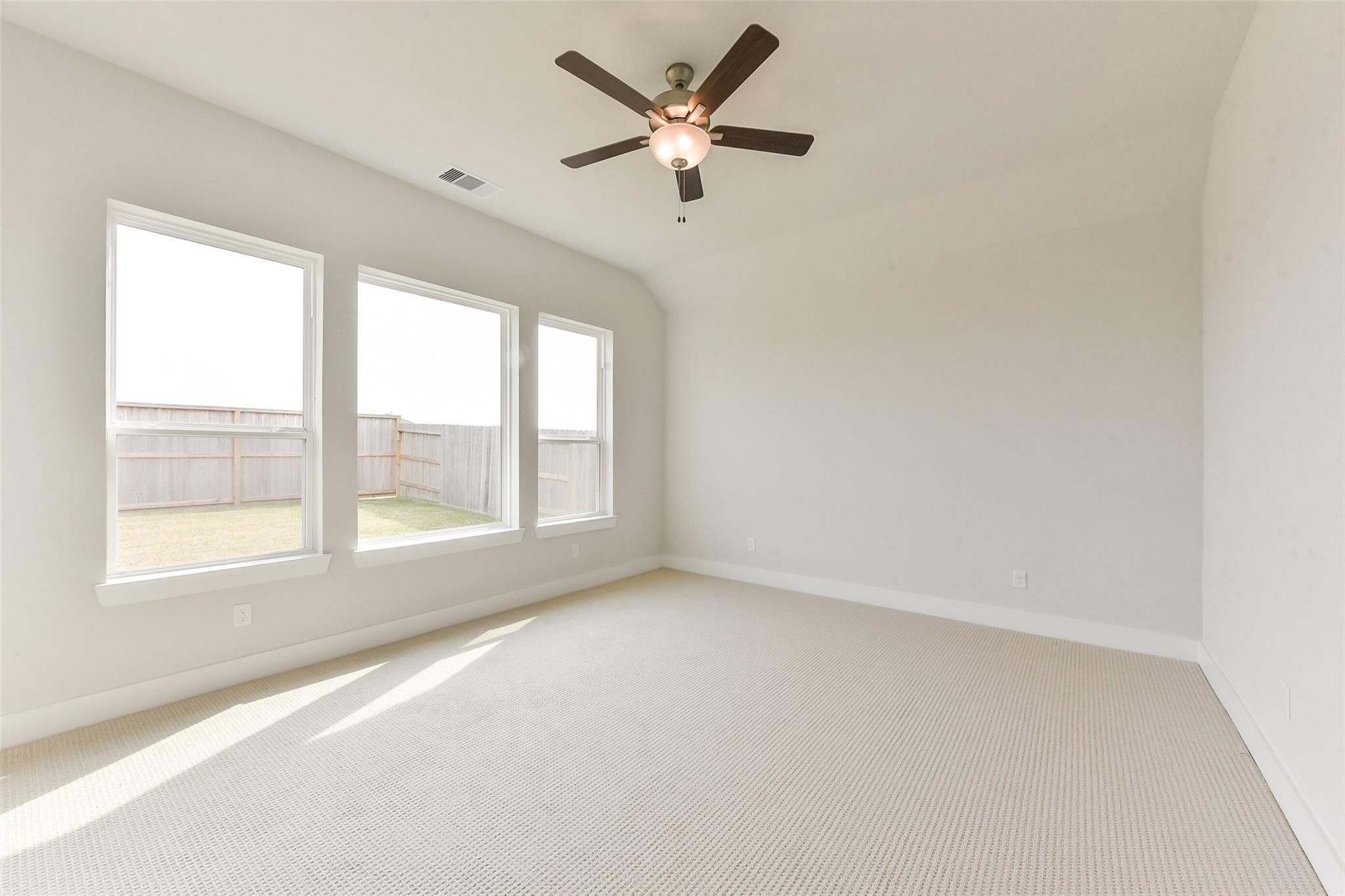 Bright empty bedroom with beige carpet, ceiling fan, and large windows overlooking fenced backyard in Davidson Homes The Edward A, Lago Mar, Texas City