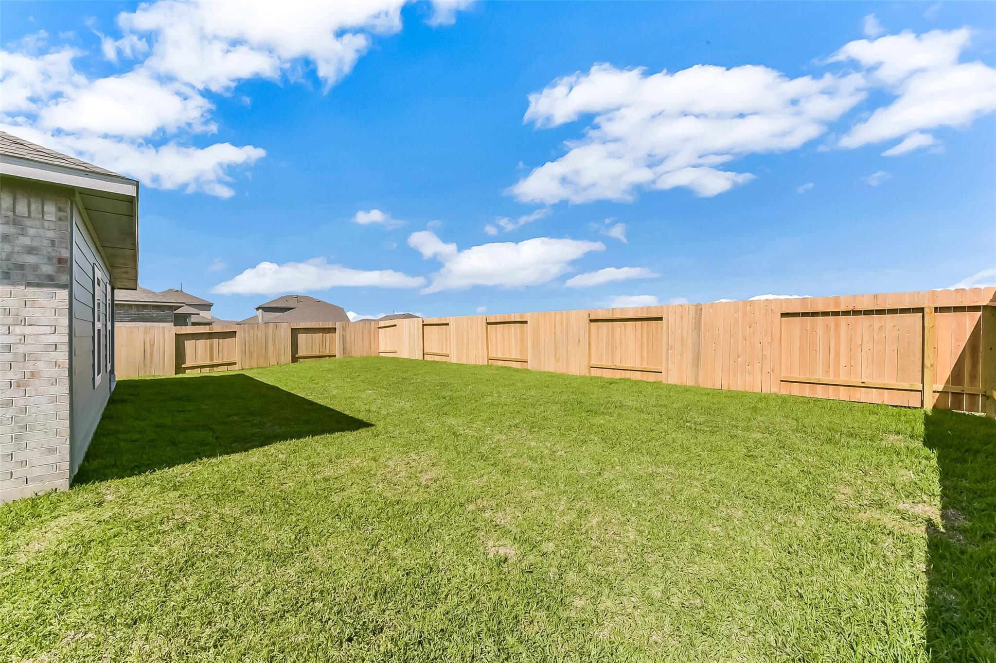 Private fenced backyard with lush green lawn, brick home exterior, and blue sky in River Ranch Meadows, Dayton, Texas