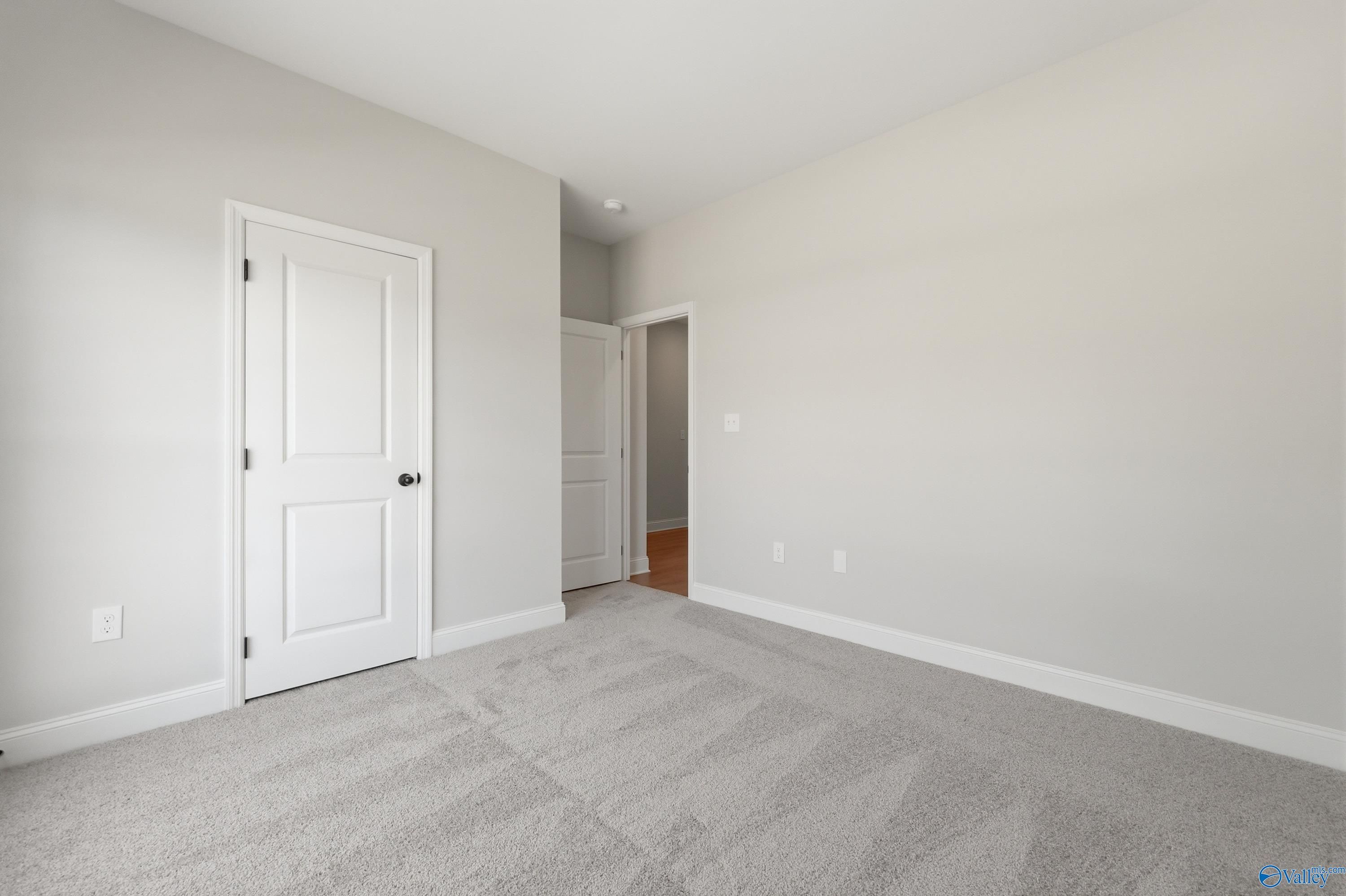 Spacious empty bedroom with light gray walls, plush gray carpet, white panel doors, and adjacent room view in Davidson Homes The Franklin, Huntsville AL