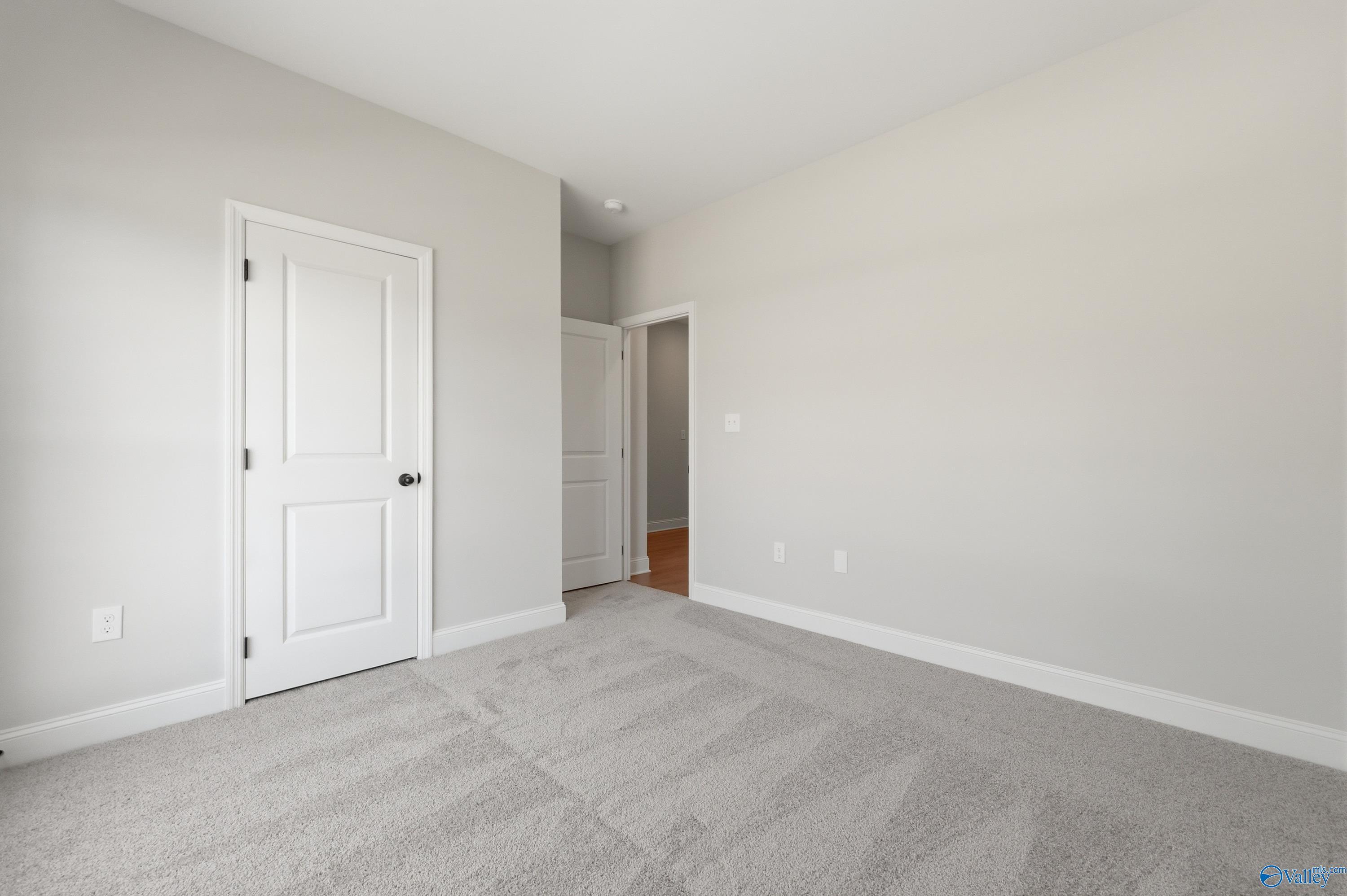 Spacious empty bedroom with light gray walls, plush gray carpet, white panel doors, and adjacent room view in Davidson Homes The Franklin, Huntsville AL