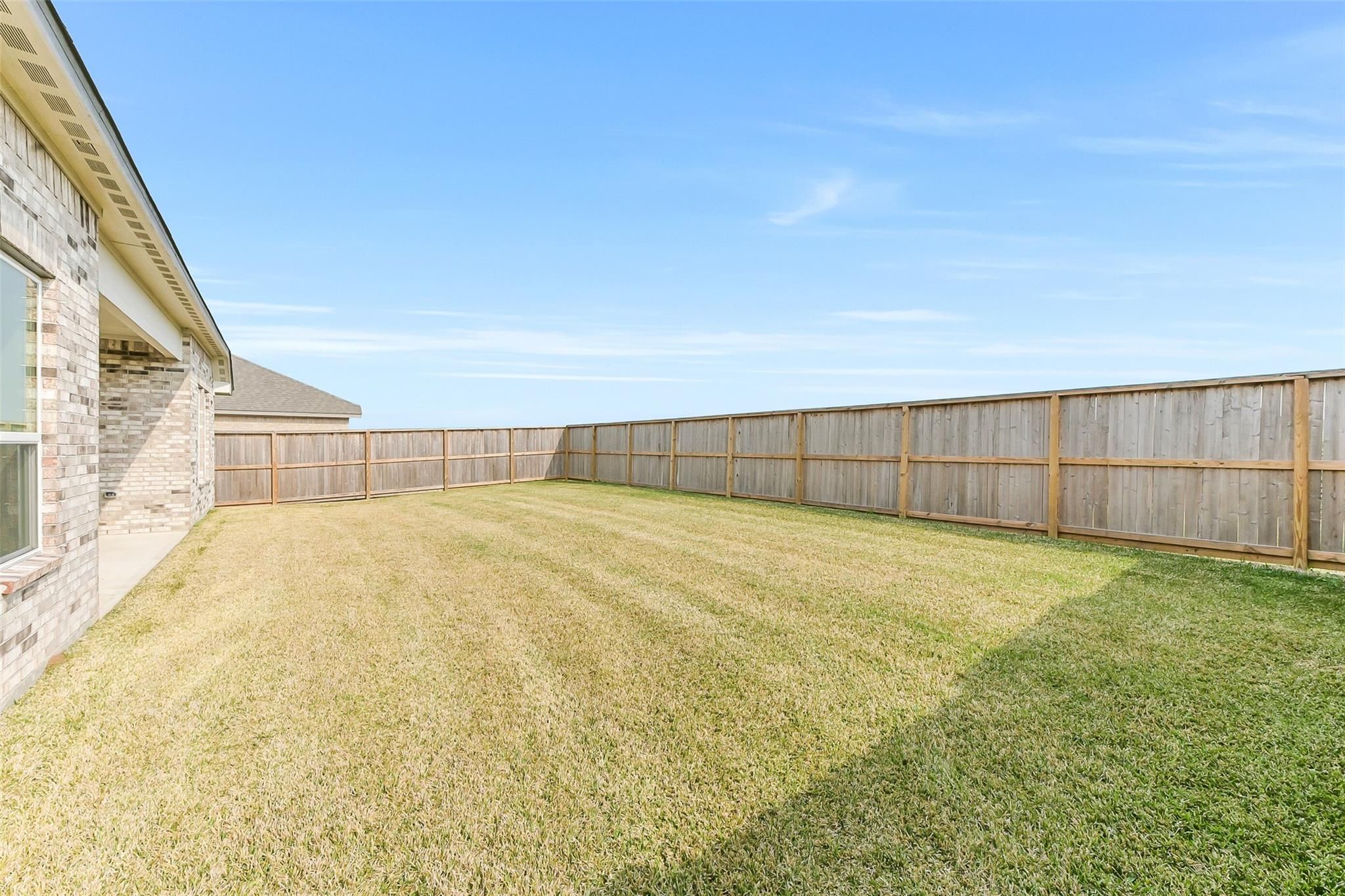Fenced backyard with patchy grass and wooden privacy fence beside brick 1-story home in Lago Mar, Texas City
