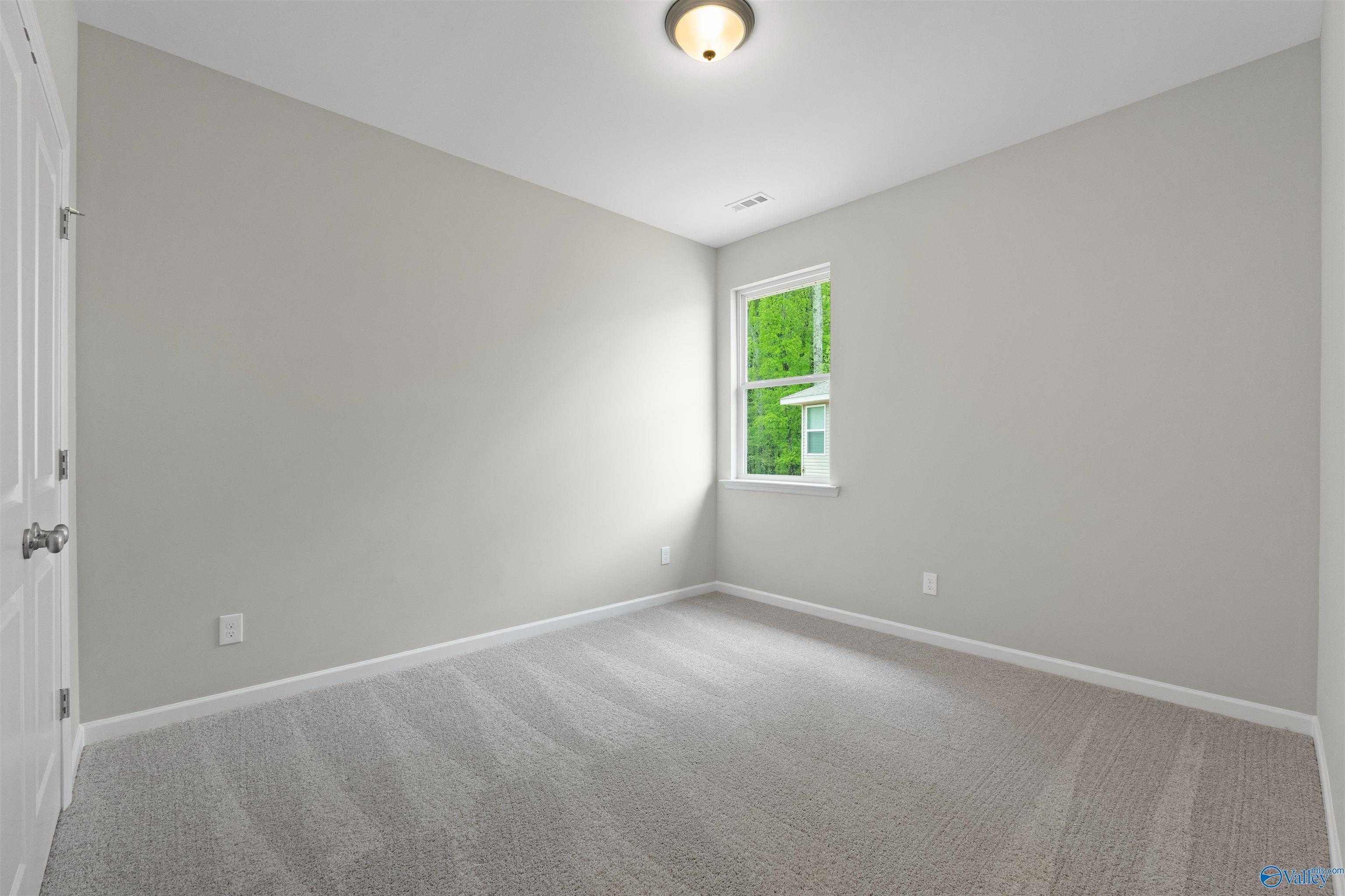 Empty secondary bedroom with light gray walls, gray carpet flooring, and window overlooking green trees in Davidson Homes The Luna, Hazel Green, Alabama