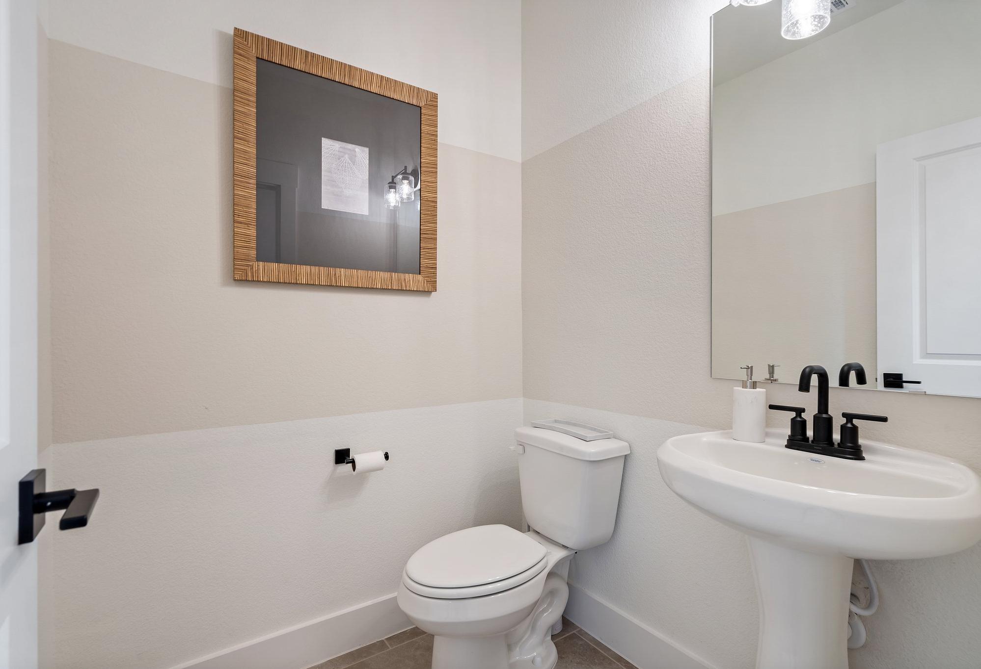 The Wake C powder room by Davidson Homes: white pedestal sink, toilet, black fixtures, beige walls, framed art in Wylie TX