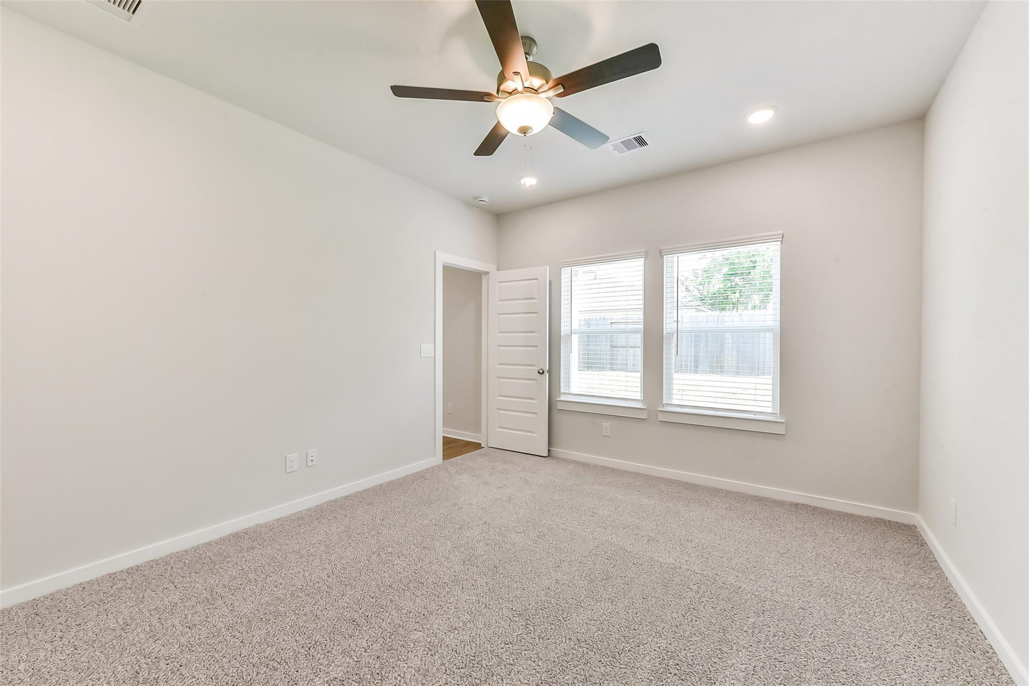 Spacious empty bedroom with beige carpet, ceiling fan, large windows, and walk-in closet door in Davidson Homes The Brazos E, Magnolia, Texas