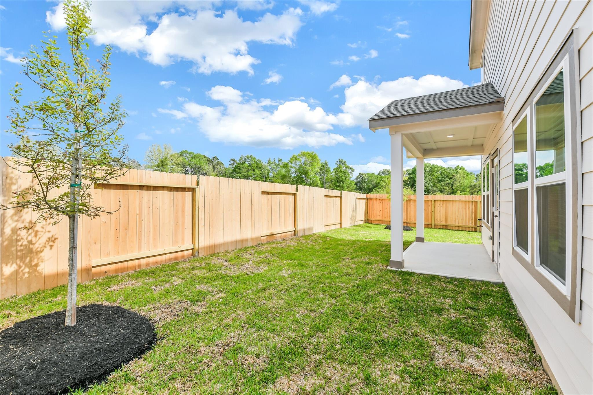 Spacious backyard with covered patio, green lawn, young tree, and wooden fence in 5-bedroom San Marcos E home, Conroe Texas