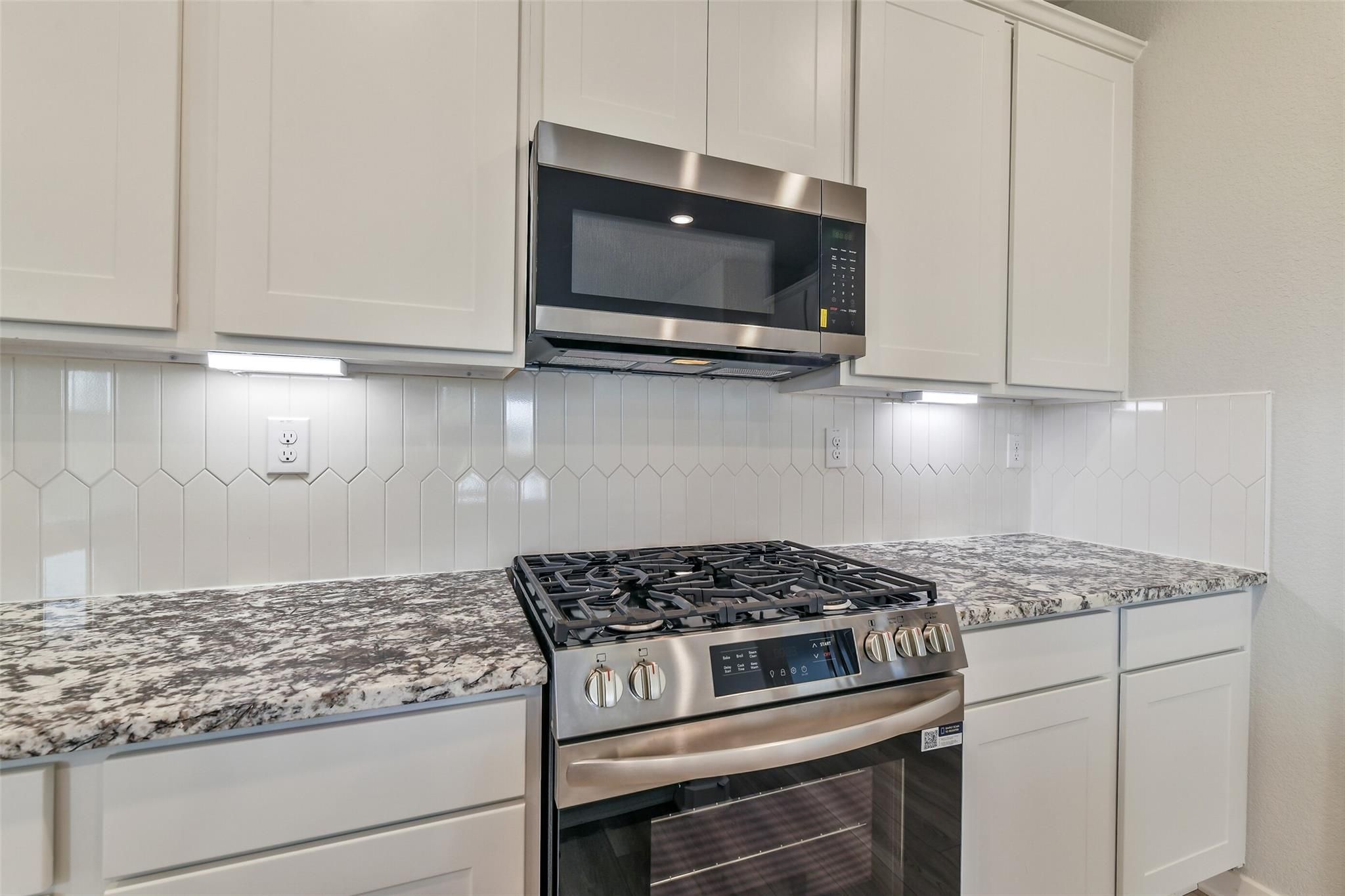 Modern white kitchen with stainless gas range, microwave, quartz counters, and hexagonal tile backsplash in Davidson Homes The Colorado G, Magnolia TX