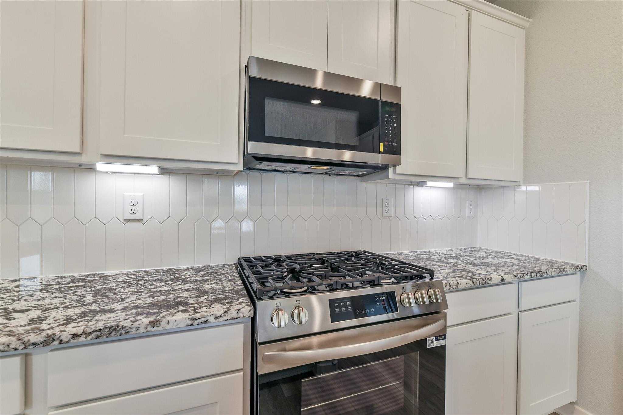 Modern white kitchen with stainless gas range, microwave, quartz counters, and hexagonal tile backsplash in Davidson Homes The Colorado G, Magnolia TX