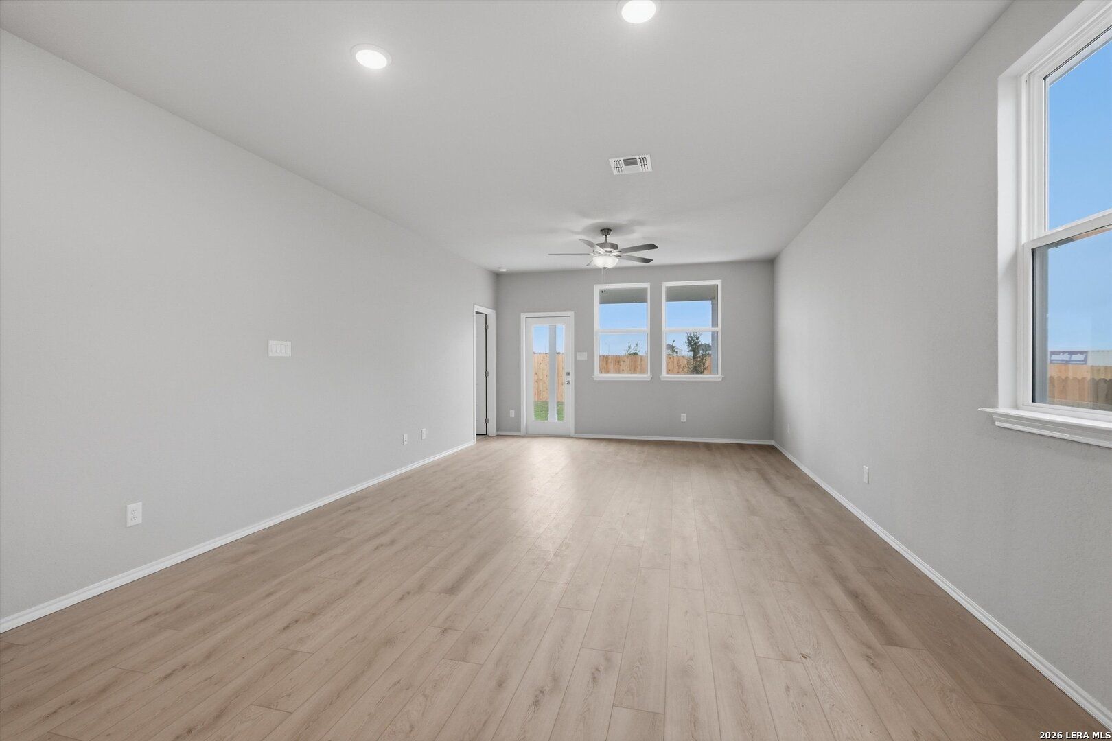 Spacious empty living room with gray walls, wood laminate floors, ceiling fan, and large windows to backyard in The Frio B, San Antonio