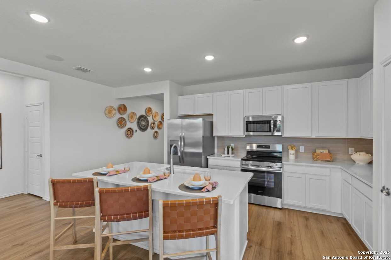 Modern white kitchen with stainless steel appliances, large island seating, and woven wall decor in Davidson Homes The Daphne J, Elmendorf, TX