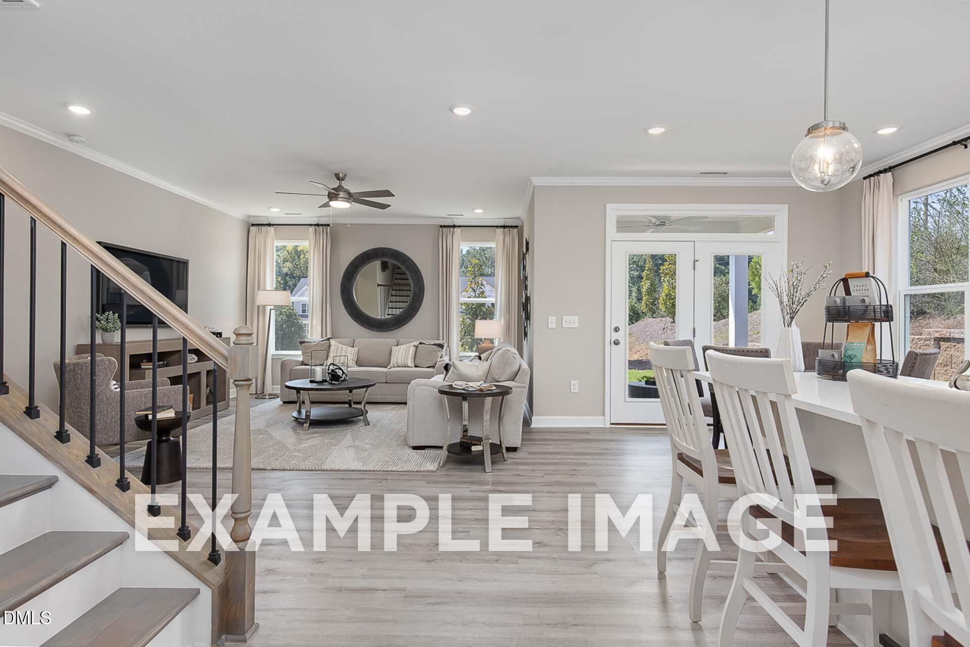 Open-concept living and dining area with beige sofa, white chairs, mounted TV, round mirror, and staircase in The Ash B 3-bedroom home, Lillington, NC
