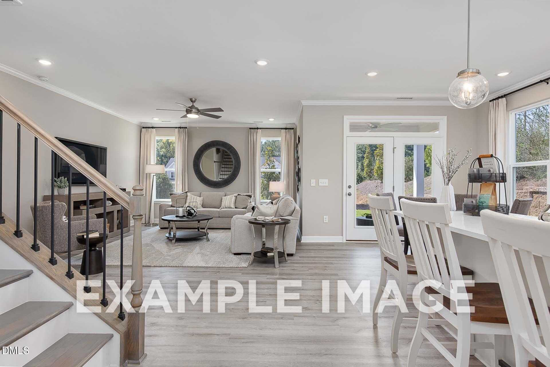 Open-concept living and dining area with beige sofa, white chairs, mounted TV, round mirror, and staircase in The Ash B 3-bedroom home, Lillington, NC