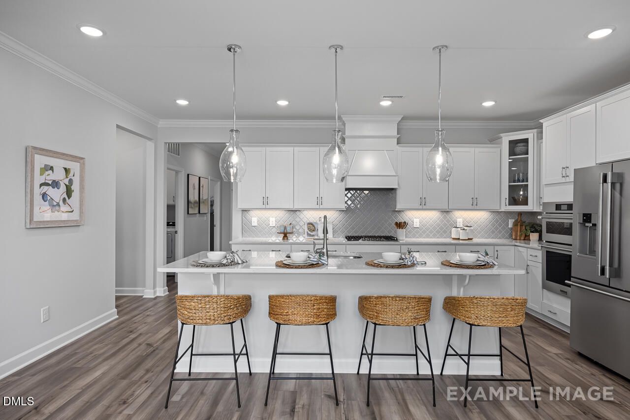Modern white kitchen with large island, woven bar stools, stainless appliances, and pendant lights in The Magnolia D, Angier, NC