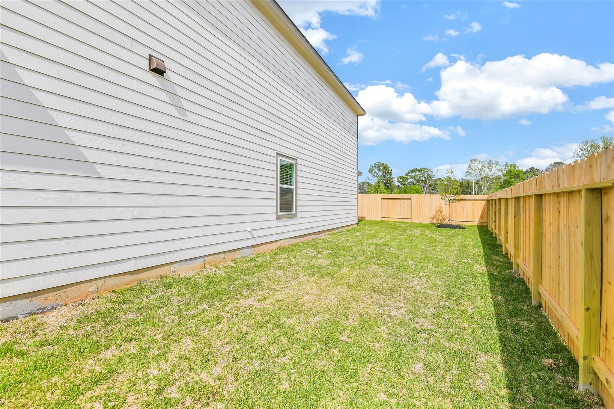 White-sided two-story home exterior with side window, lush green lawn, and wooden privacy fence in Caney Creek Place, Conroe, Texas