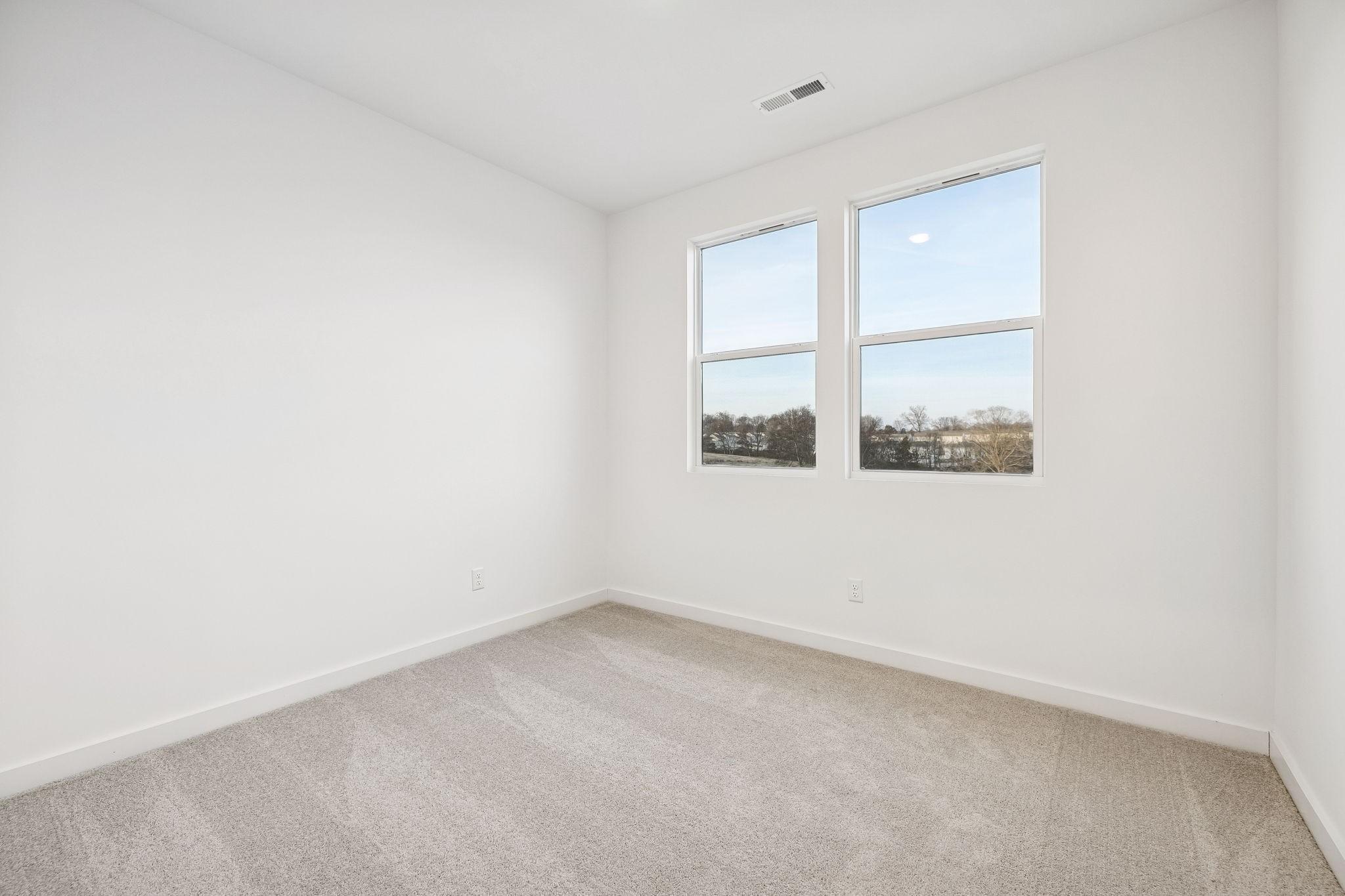 Bright empty bedroom with large double windows overlooking trees, beige carpet, white walls in The Willow, Davidson Homes, White House, TN