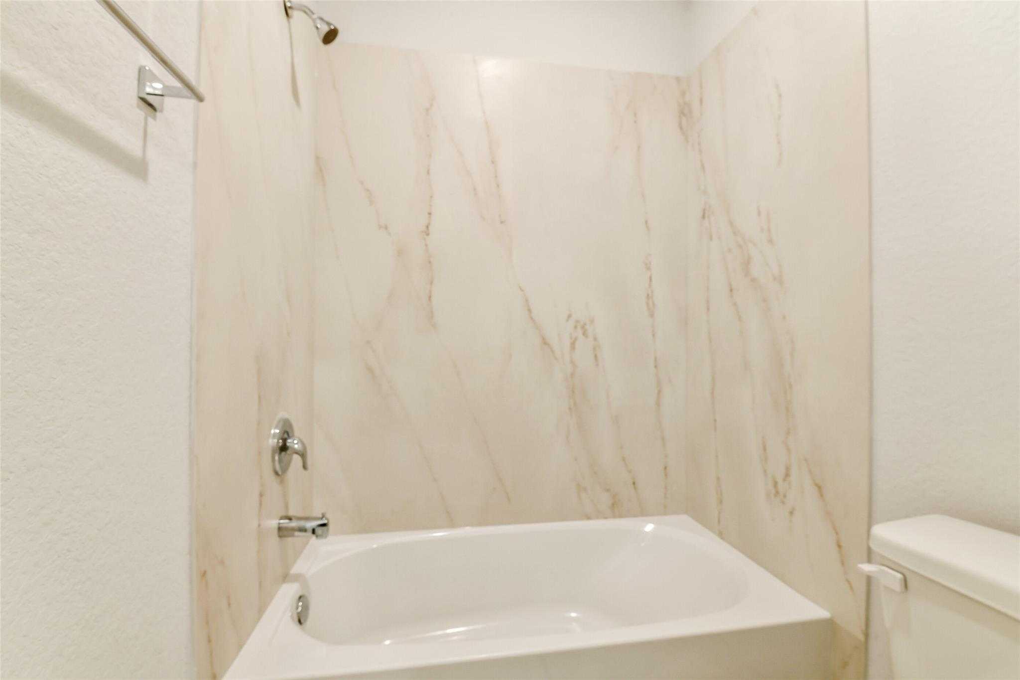 Elegant bathroom featuring marble-look beige tiled walls, deep soaking tub with shower, and adjacent toilet in Davidson Homes The Colorado F, Cleveland, Texas