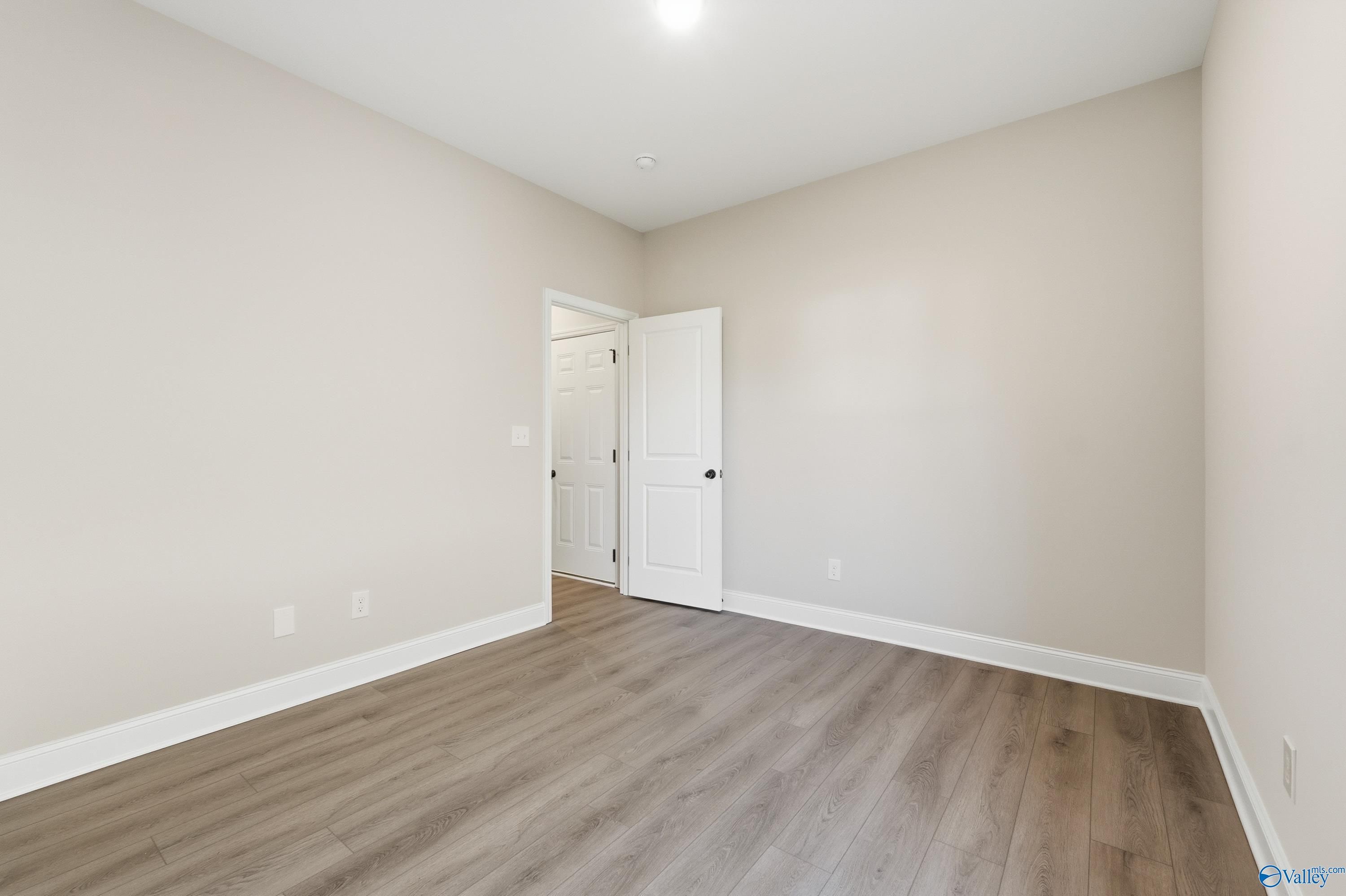 Spacious empty bedroom with beige walls, light wood flooring, and white door in Davidson Homes The Daphne D, Arab, Alabama