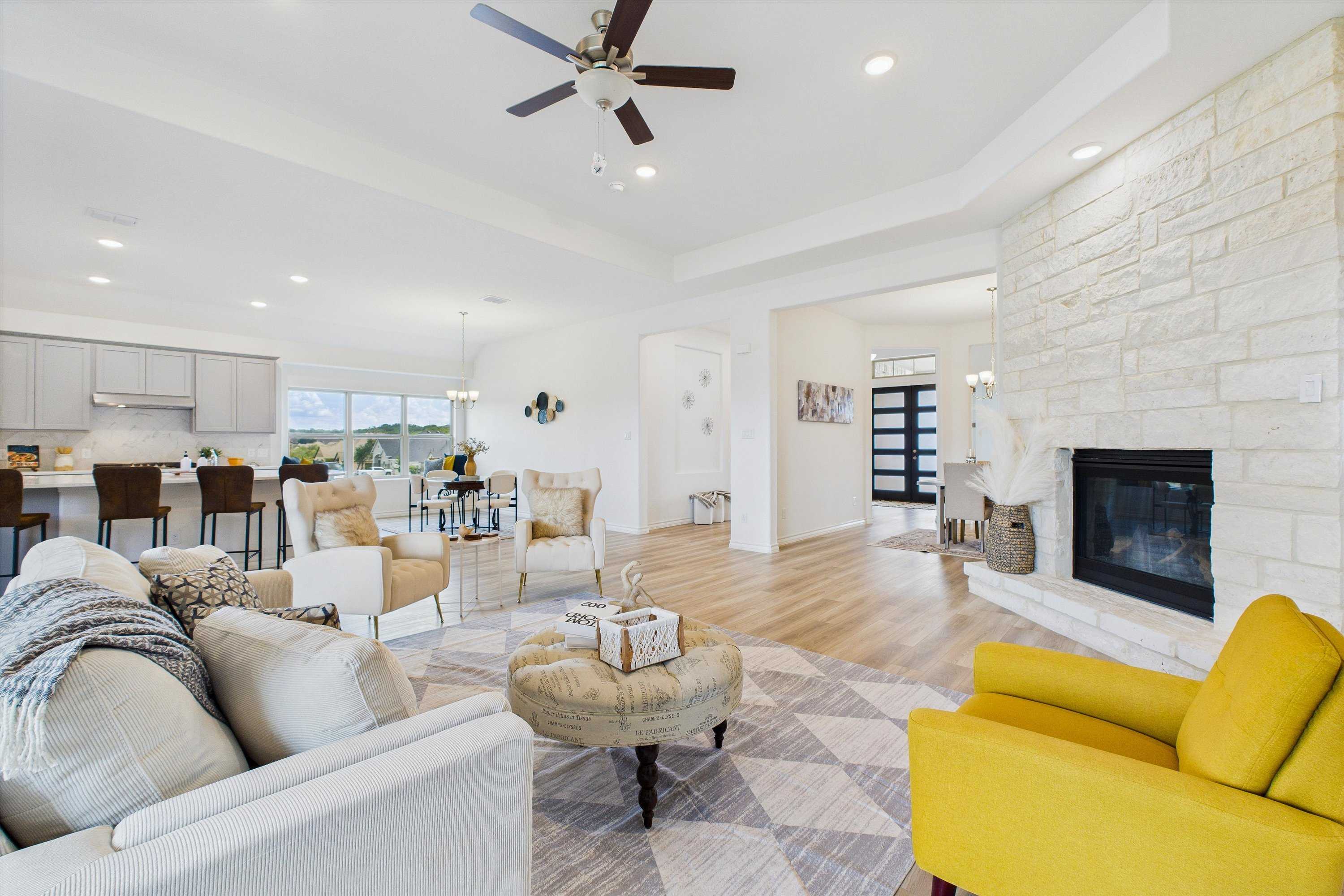 Open-concept living room with stone fireplace, white sofas, yellow chair, and adjacent kitchen bar in Davidson Homes The Summerlin C, Castroville, Texas