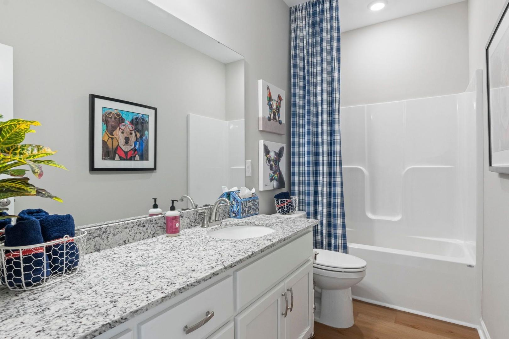 Bright secondary bathroom in The Phoenix A featuring granite vanity, white cabinets, bathtub, blue shower curtain, and dog-themed art