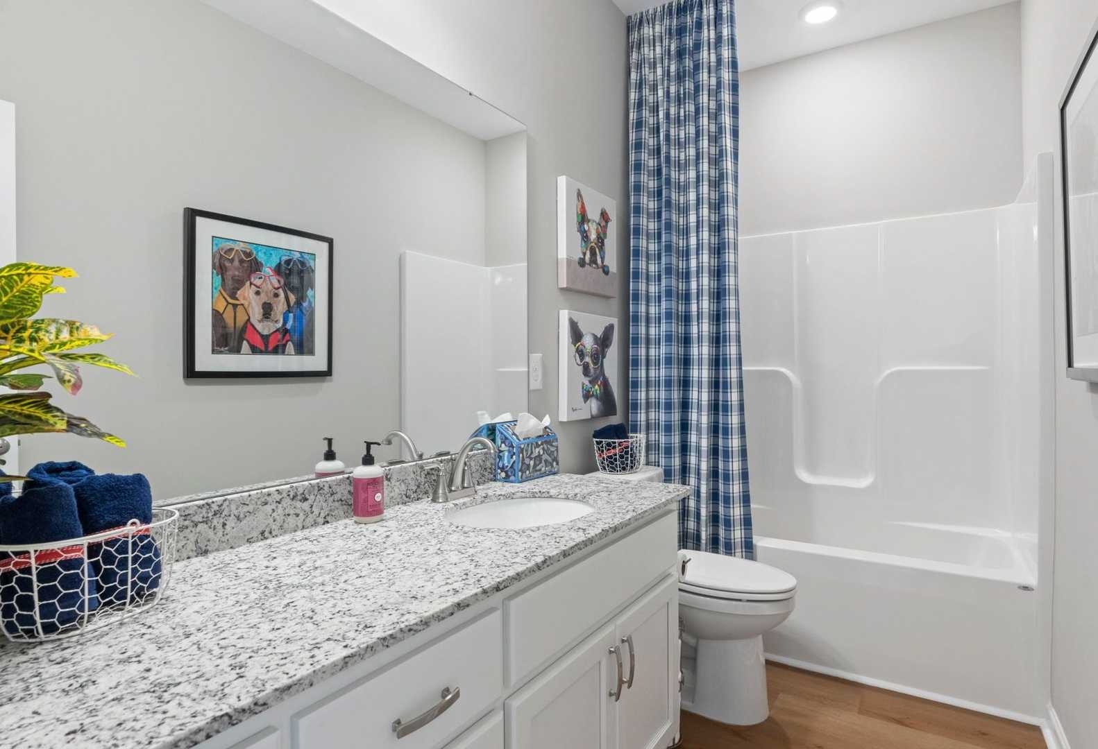Bright secondary bathroom in The Phoenix A featuring granite vanity, white cabinets, bathtub, blue shower curtain, and dog-themed art