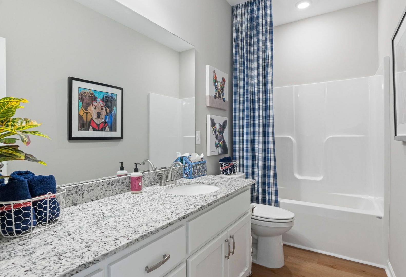 Bright secondary bathroom in The Phoenix A featuring granite vanity, white cabinets, bathtub, blue shower curtain, and dog-themed art