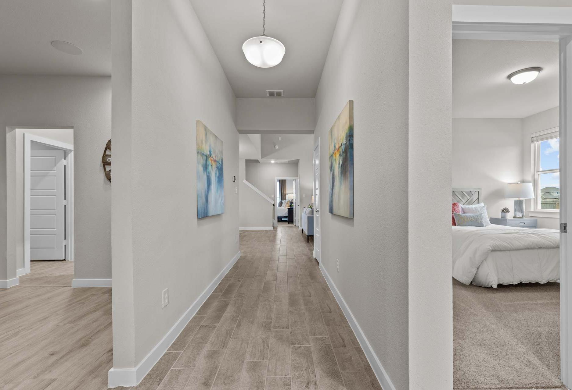 Spacious gray hallway with hardwood floors, abstract wall art, and open doorway to airy master bedroom in The Collin A, Seguin TX
