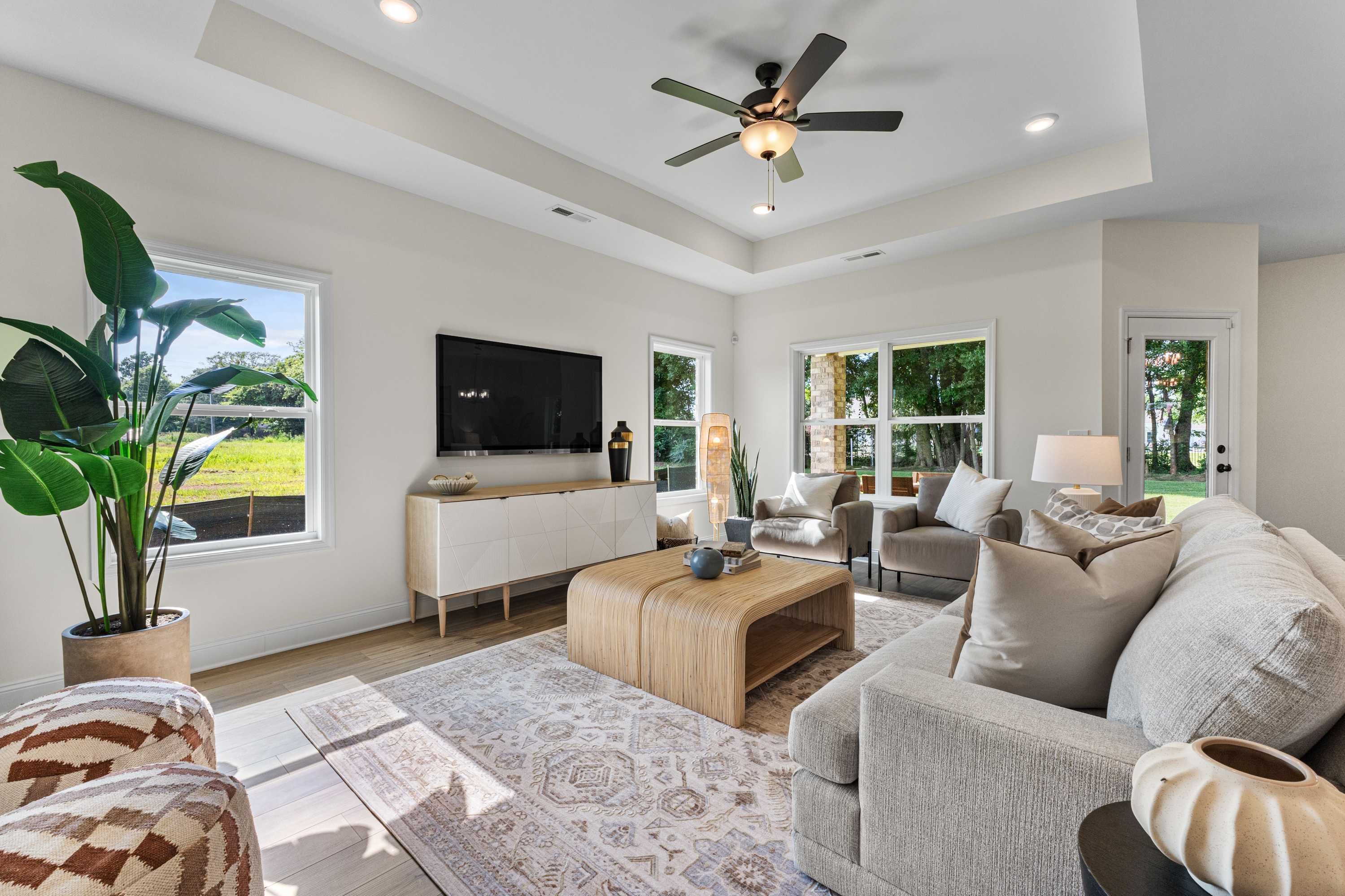 Spacious living room at Lynn Meadows in Meridianville AL with beige sectional sofa, mounted TV, ceiling fan, and large windows overlooking greenery