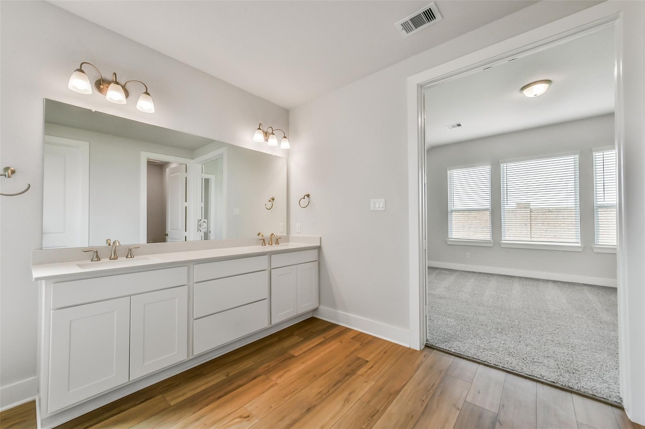 Elegant double vanity master bathroom with white shaker cabinets, gold faucets, large mirror, and adjacent carpeted bedroom in Davidson Homes The Victoria A, Lago Mar, Texas